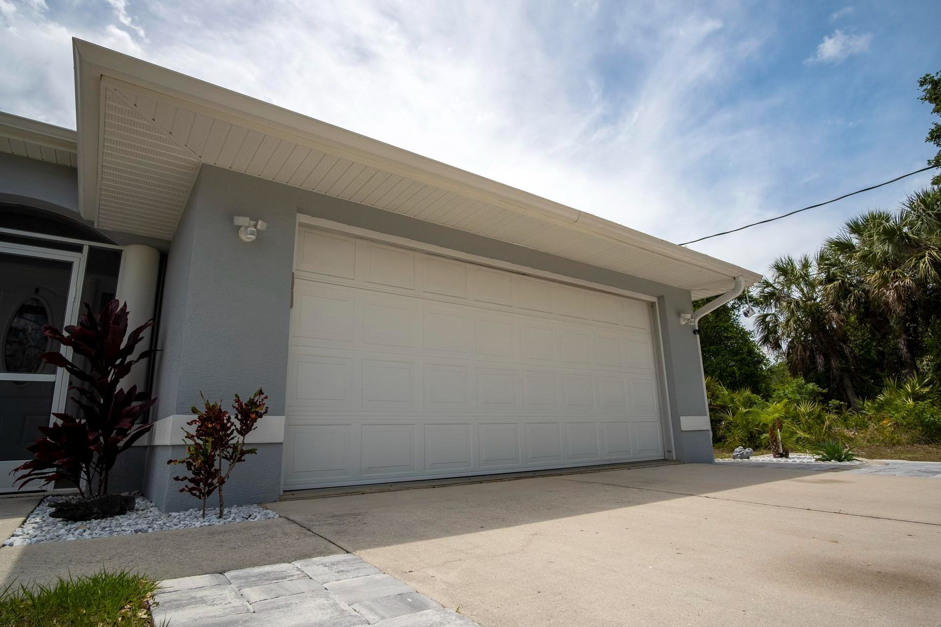 White garage door on a gray stucco house with a concrete driveway, plants, and a blue sky.