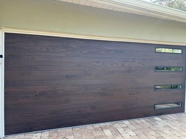 Dark brown garage door with horizontal wood grain, three rectangular windows, brick driveway.