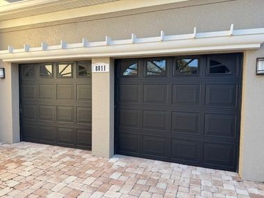Two black garage doors with windows, beige exterior, and brick driveway.