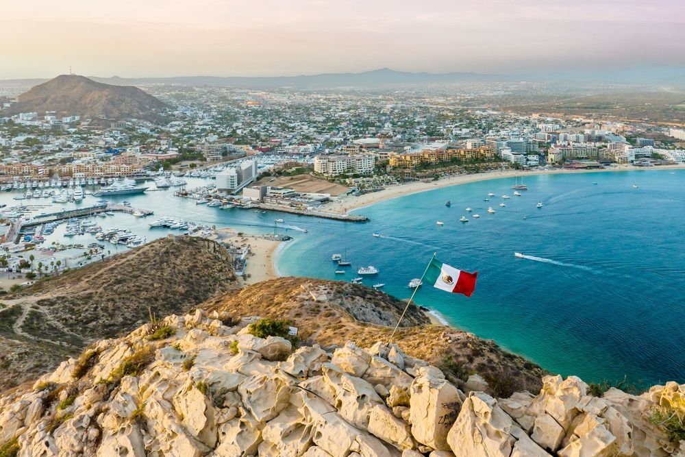 Scenic coastal view of Cabo San Lucas, Mexico, with a waving Mexican flag, city, boats, and turquoise water.