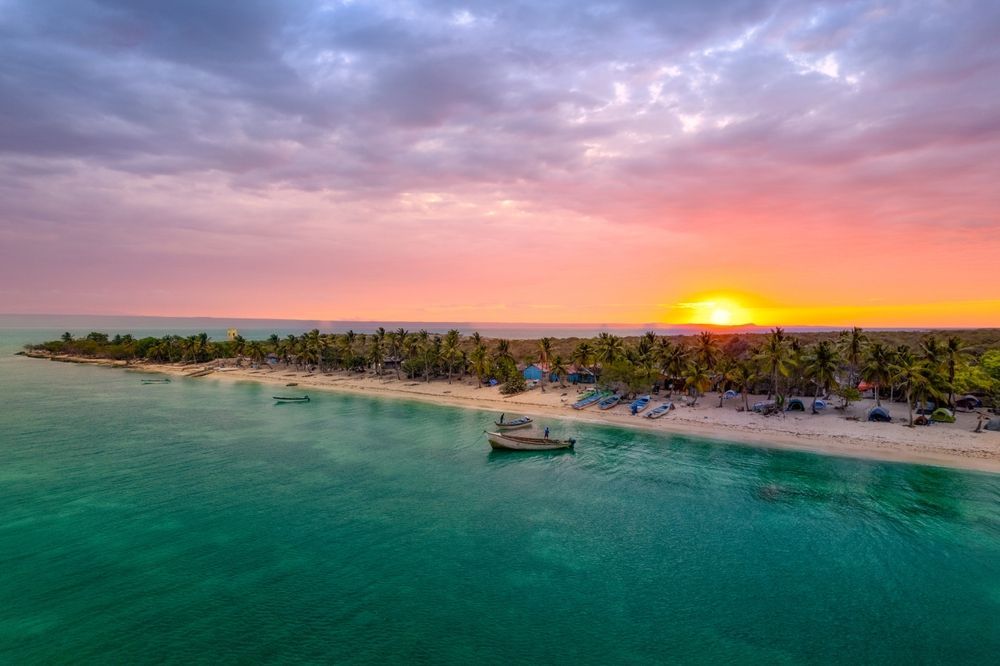 Tropical beach at sunset with palm trees, turquoise water, and a boat. Colorful sky.