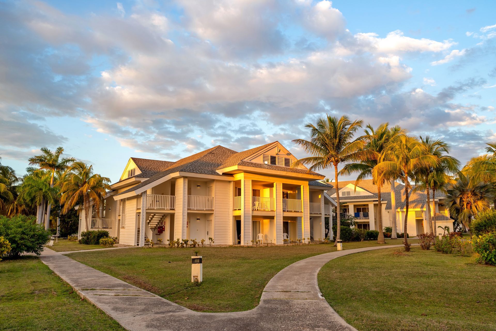 Large white building with balconies and palm trees, pathways on grassy lawn, under a cloudy sky.