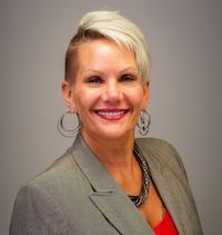 A woman in a blue shirt and earrings is leaning against a wall.