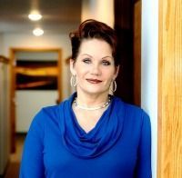 A woman in a blue shirt and earrings is leaning against a wall.