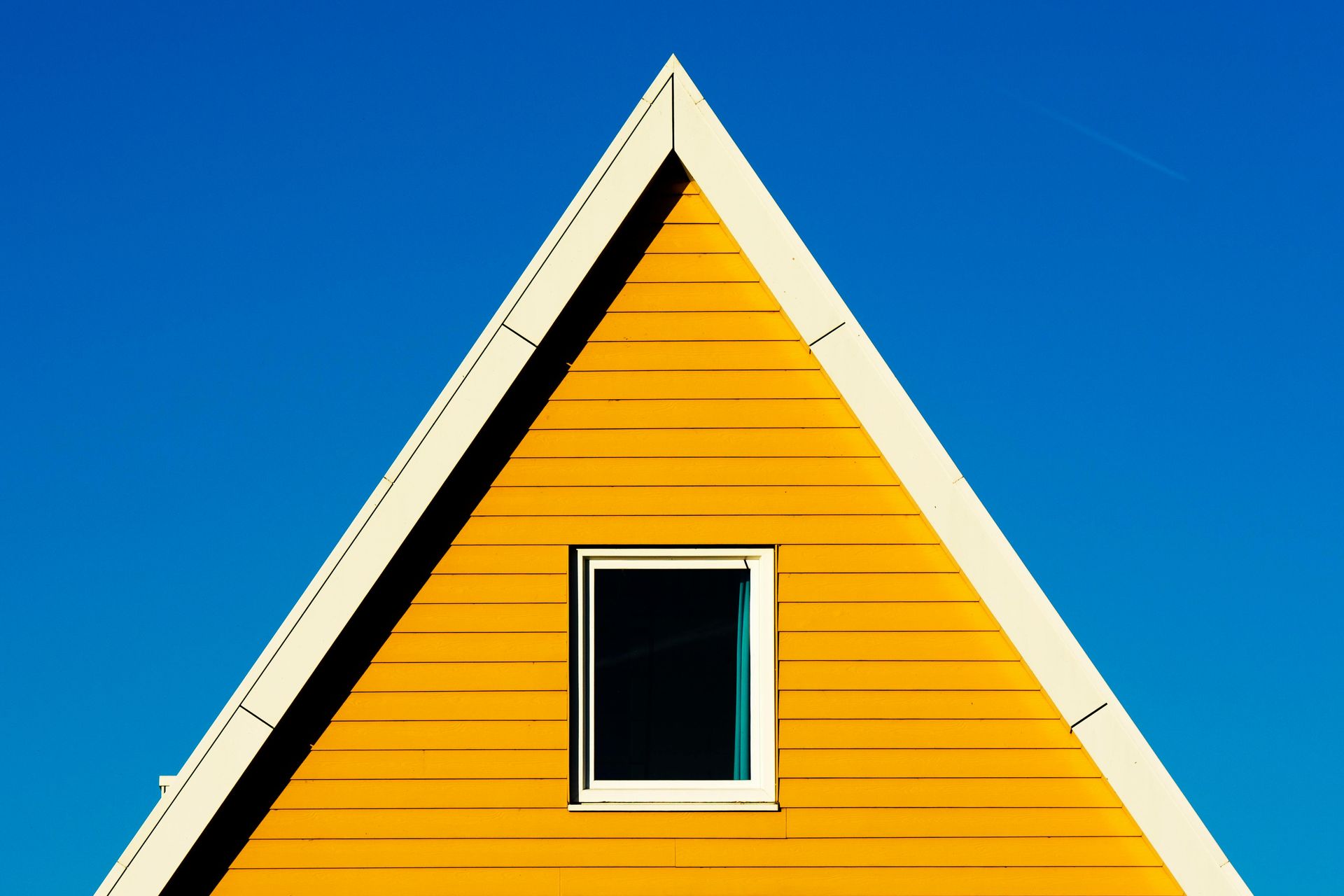 Yellow triangular building facade with white trim, small window, bright blue sky.