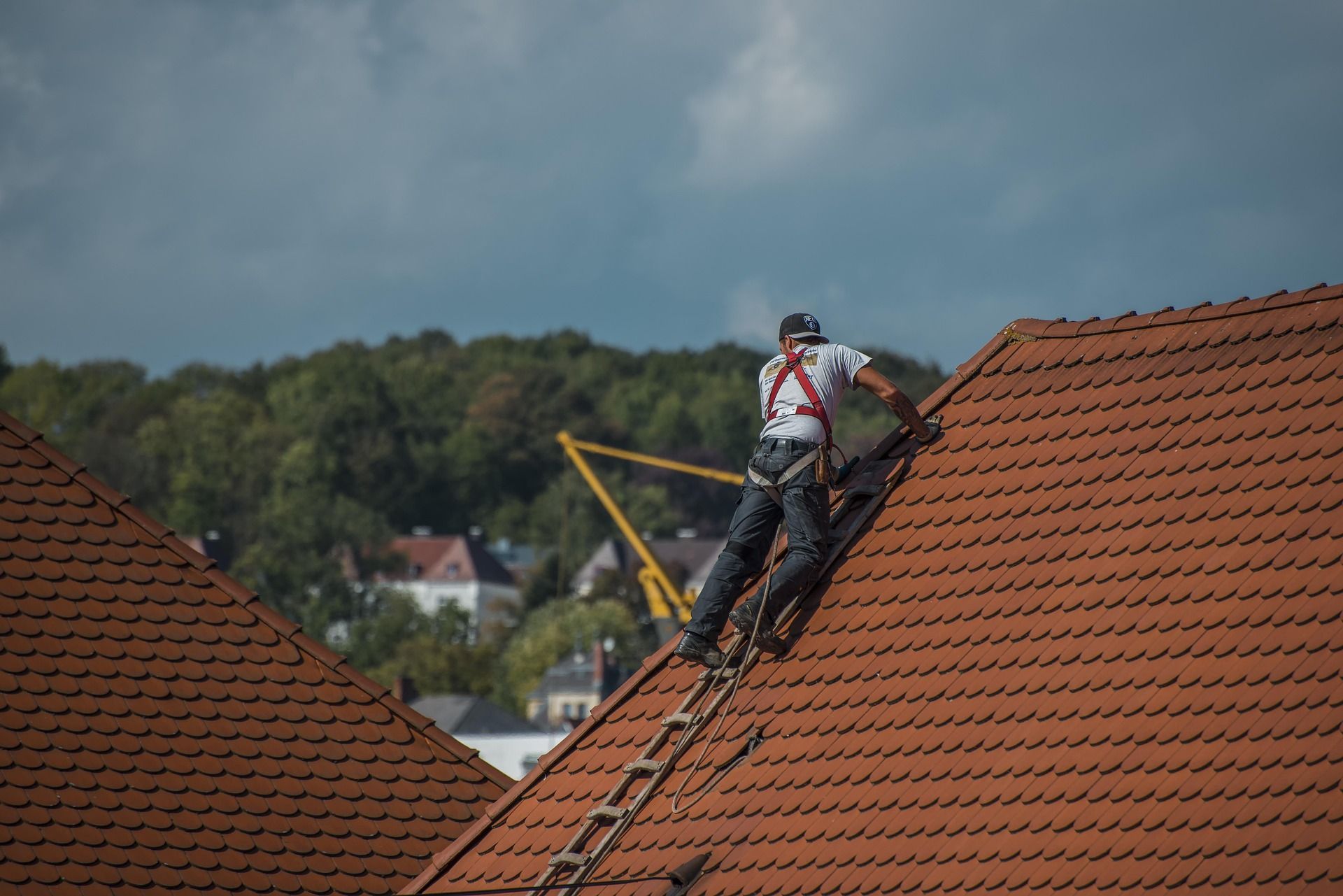 Roofer on a ladder, working on a red-tiled roof, wearing safety harness.