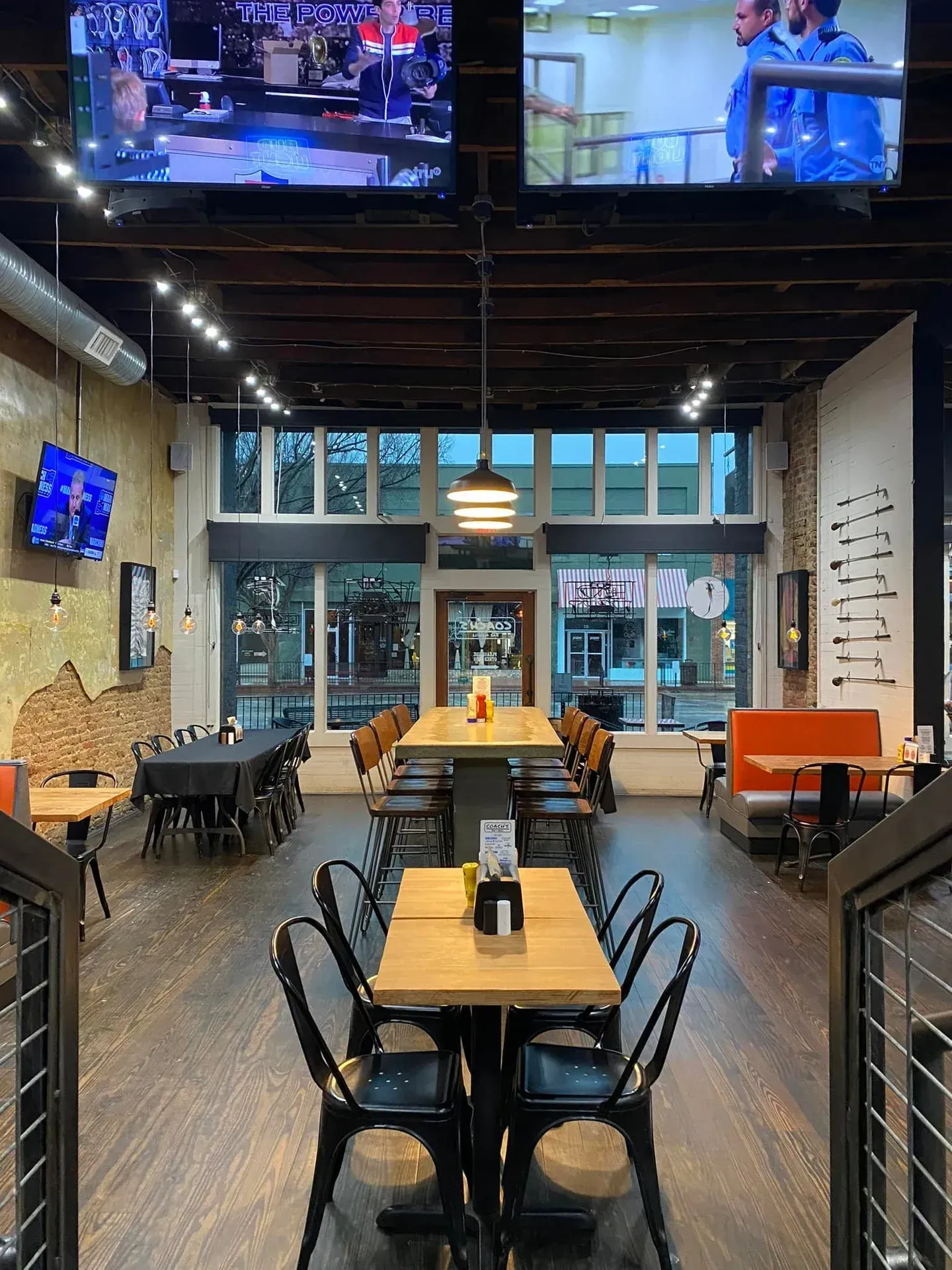 Restaurant interior with wooden tables and black chairs; TVs on the walls; exposed brick and a long communal table.