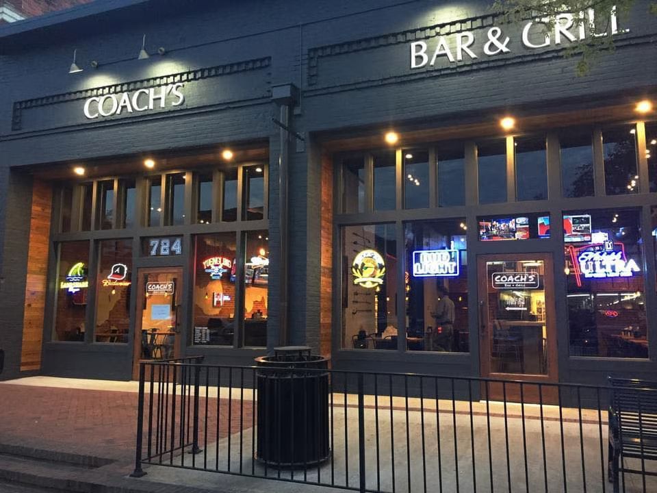 Coach's Bar & Grill storefront with large windows, lit signs, and an outdoor railing.