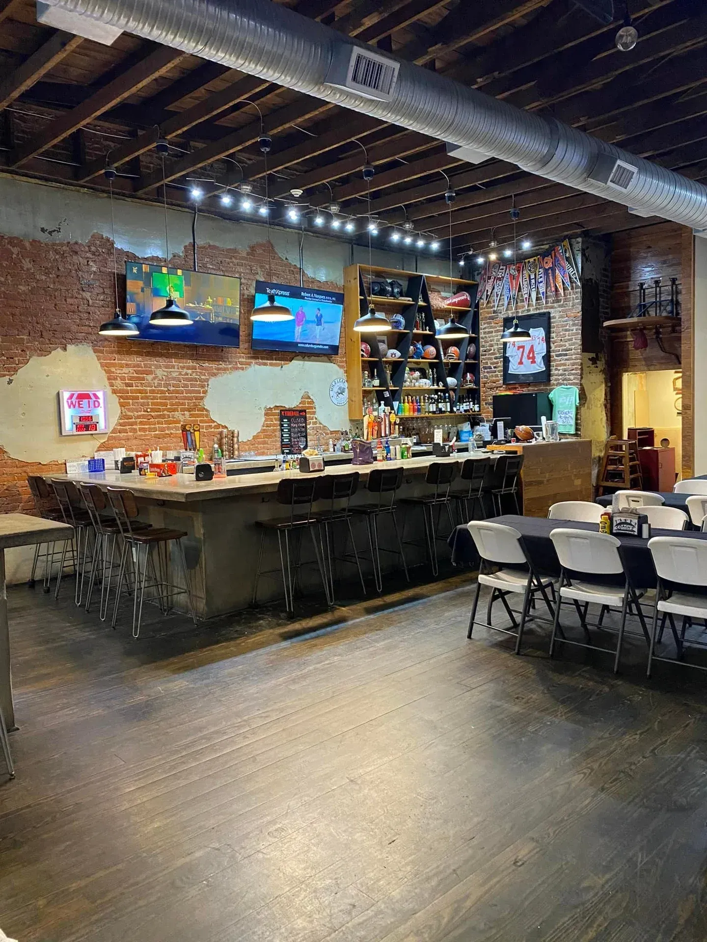 Interior view of a bar with a brick wall, wooden floor, bar stools, and TVs. Tables and chairs on the right.