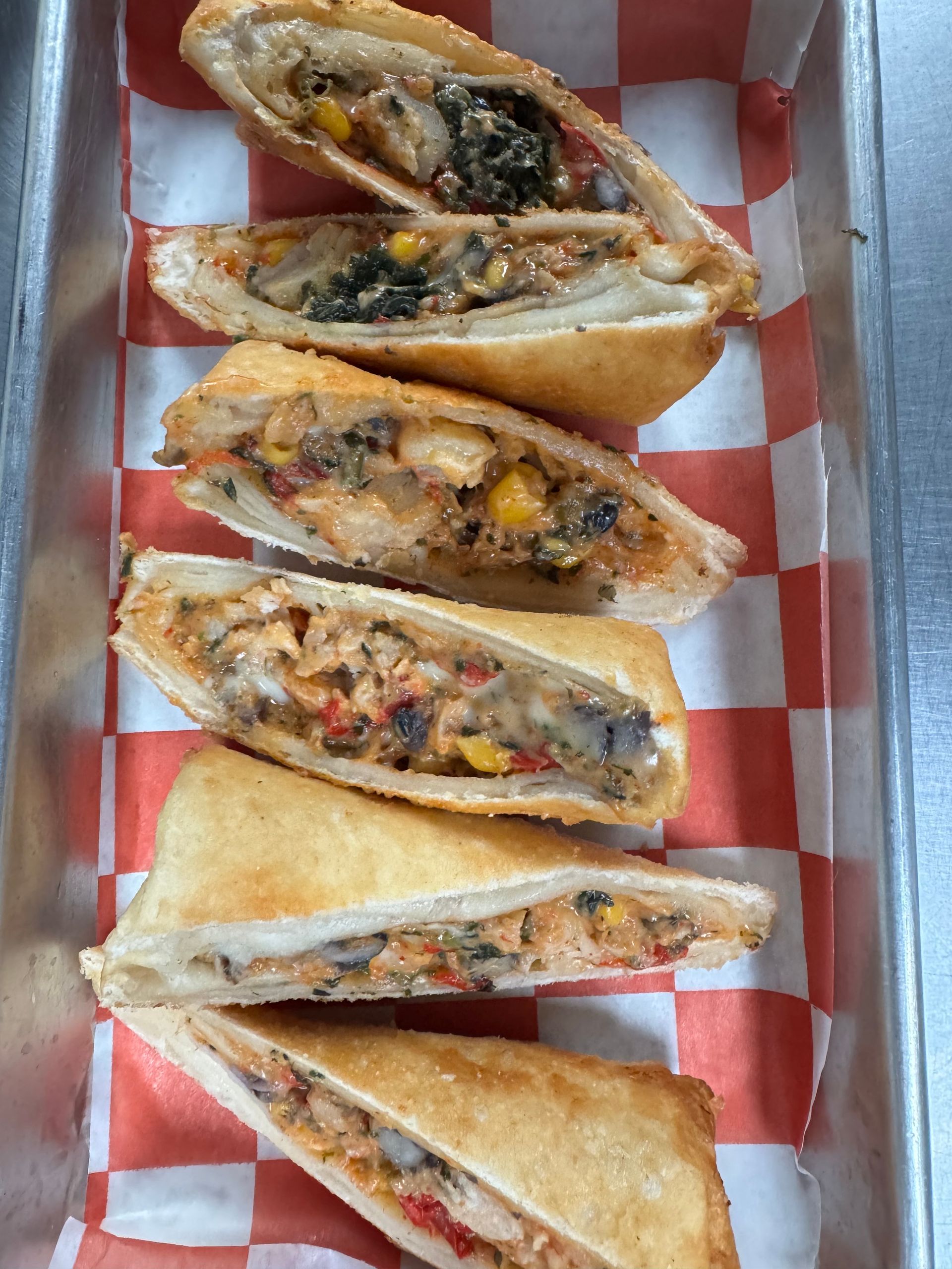 Six fried food pastries filled with vegetables and possibly meat, on a checkered red-and-white paper in a metal container.