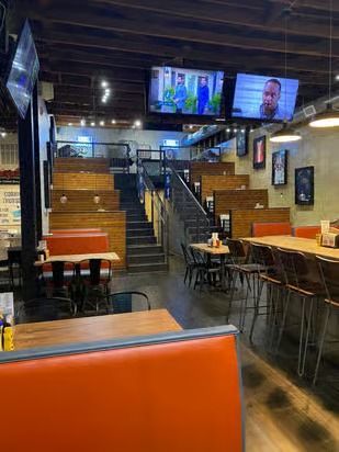 Restaurant interior with tiered seating, tables, TVs, and a staircase. Orange booths and metal chairs.