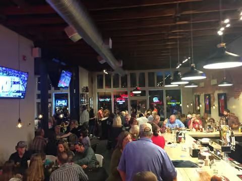 Busy bar scene with patrons, bar, tables, overhead lights, TVs, and large windows.
