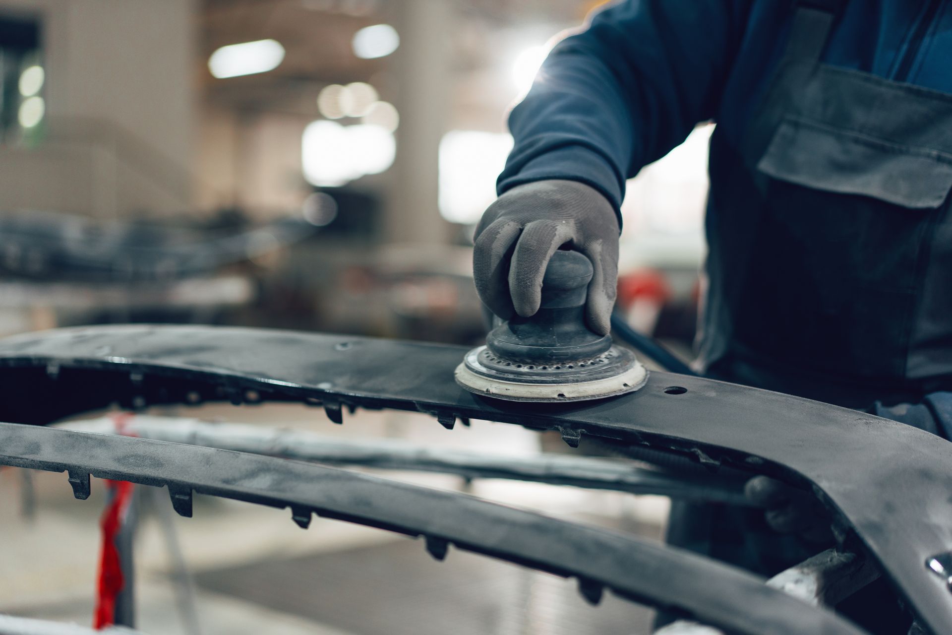 A worker in a dark uniform uses a hand-held circular sander to smooth the surface of a black car bumper in a workshop.