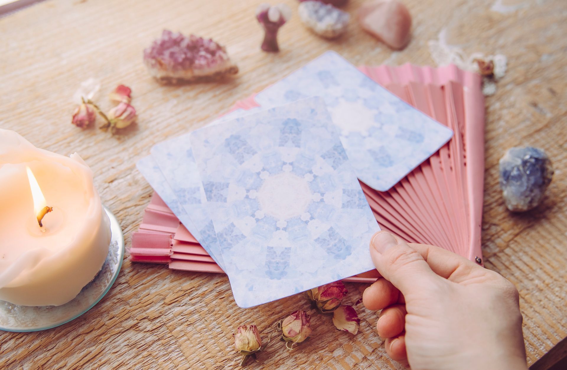 Person holding tarot cards on wooden table, with candle and crystals.