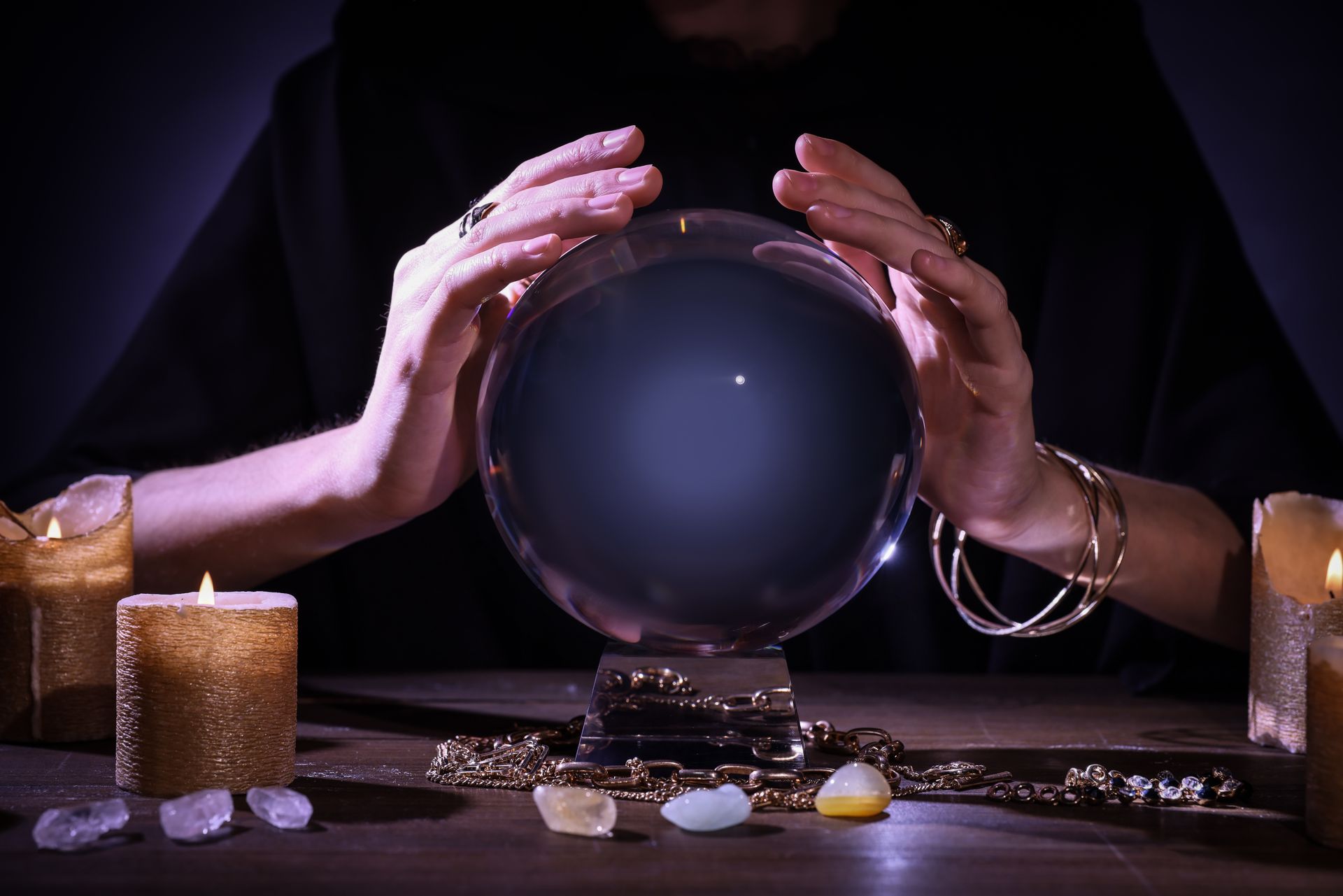 A person’s hands over a crystal ball with candles and crystals on a table. Dark background.