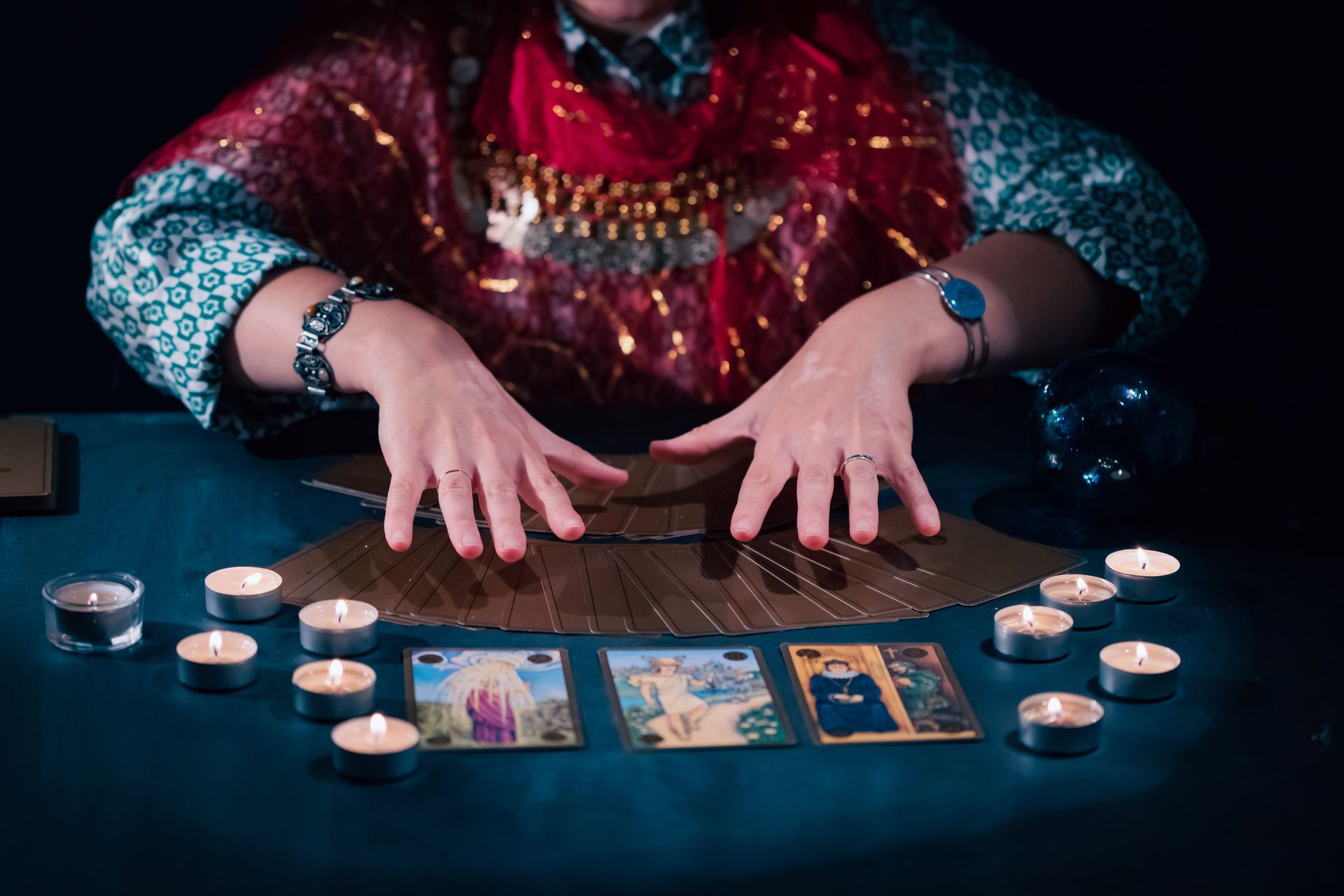 A fortune teller spreads tarot cards on a table surrounded by candles, hands visible.