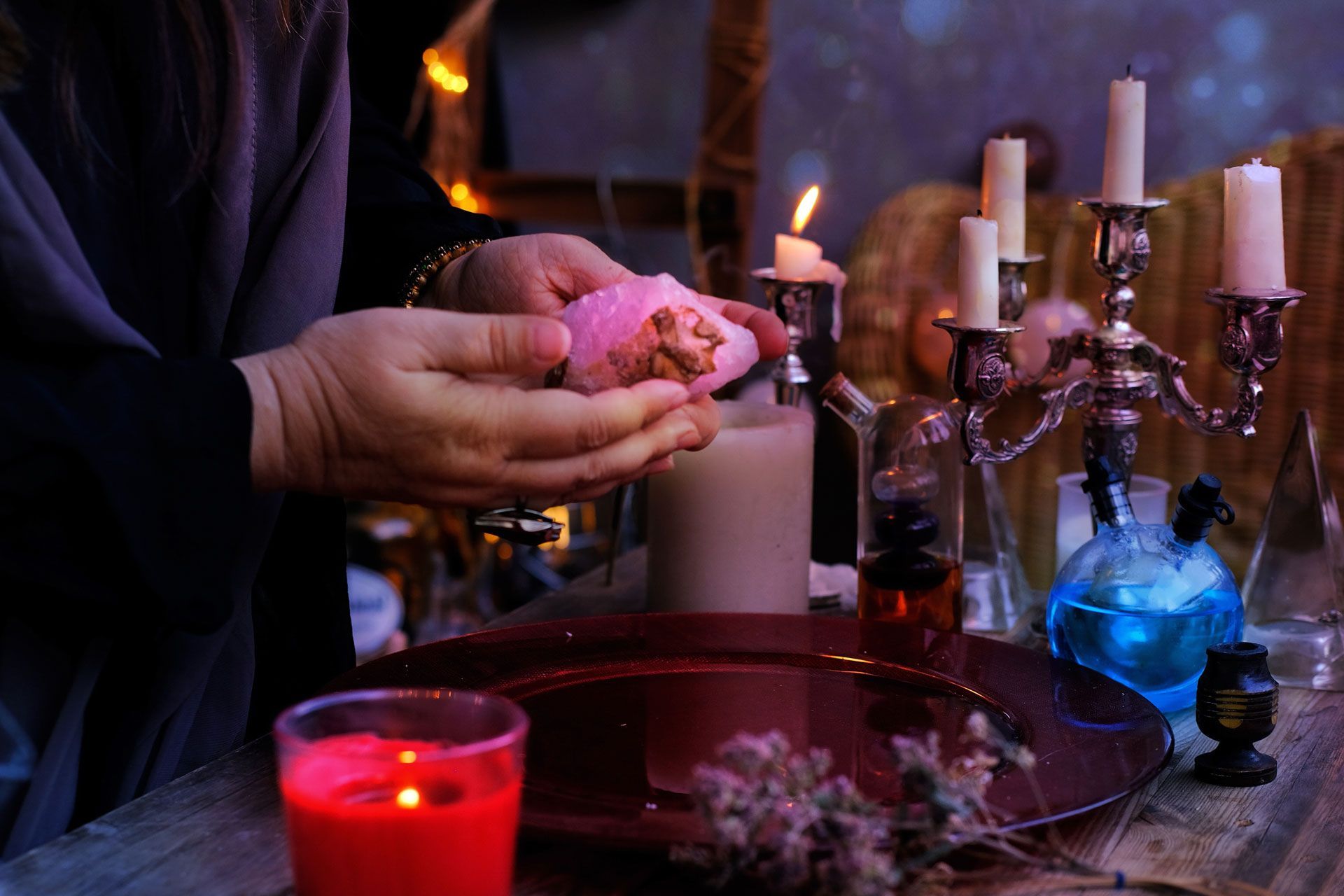 Person holding pink item over red plate with candles, potions, and other ritualistic items.