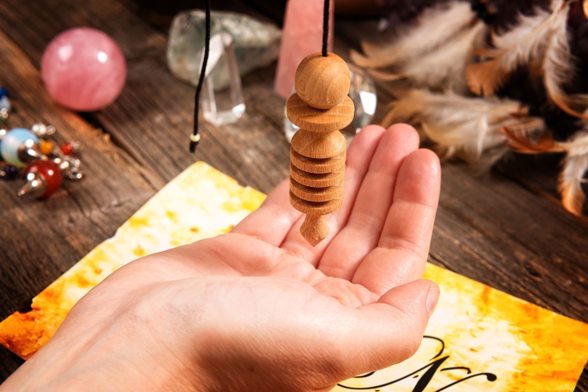 Hand holding a wooden pendulum over a map on a wooden table, crystals in the background.