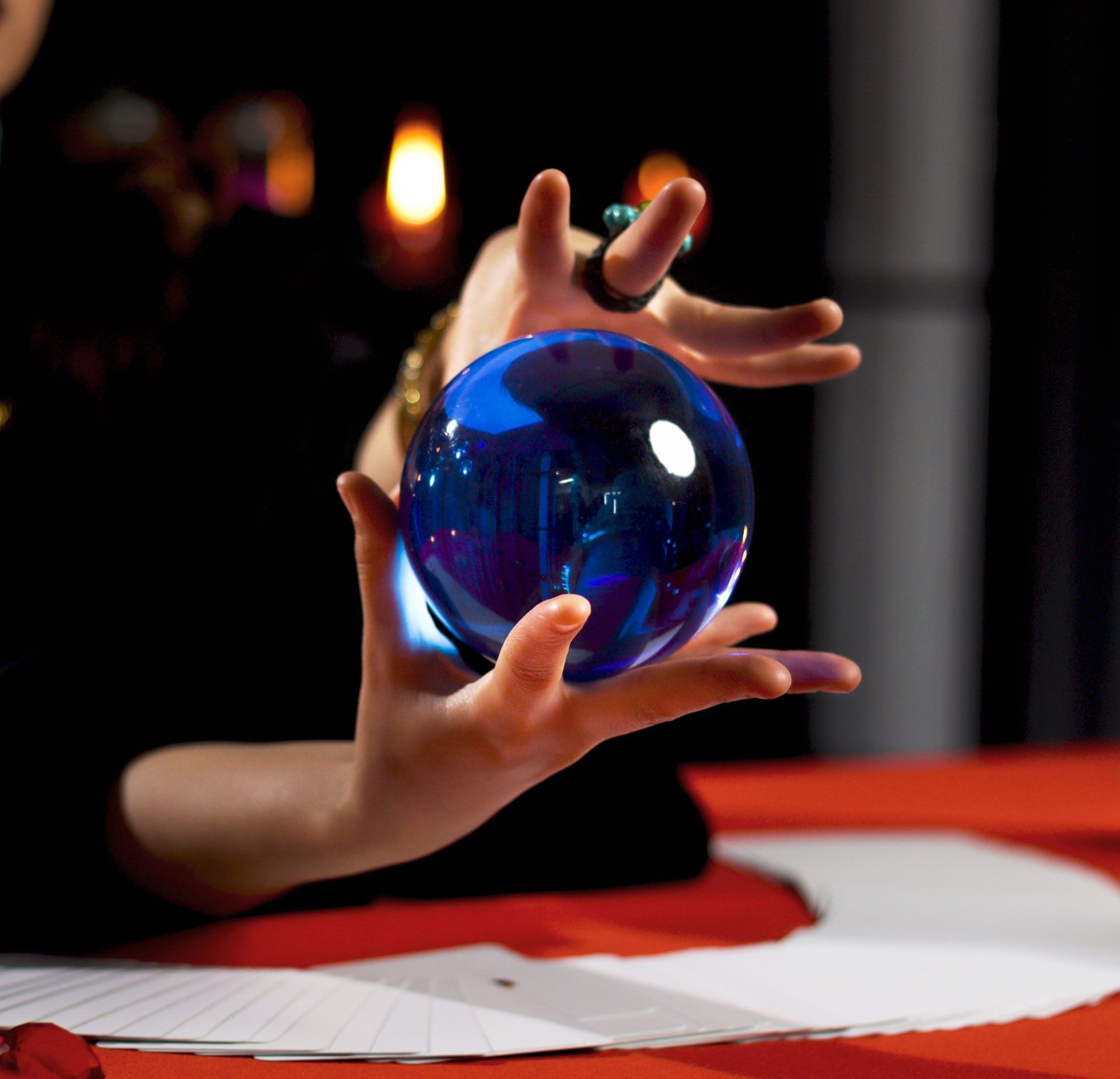 Hands holding a glowing blue crystal ball. Dark background, red tablecloth.