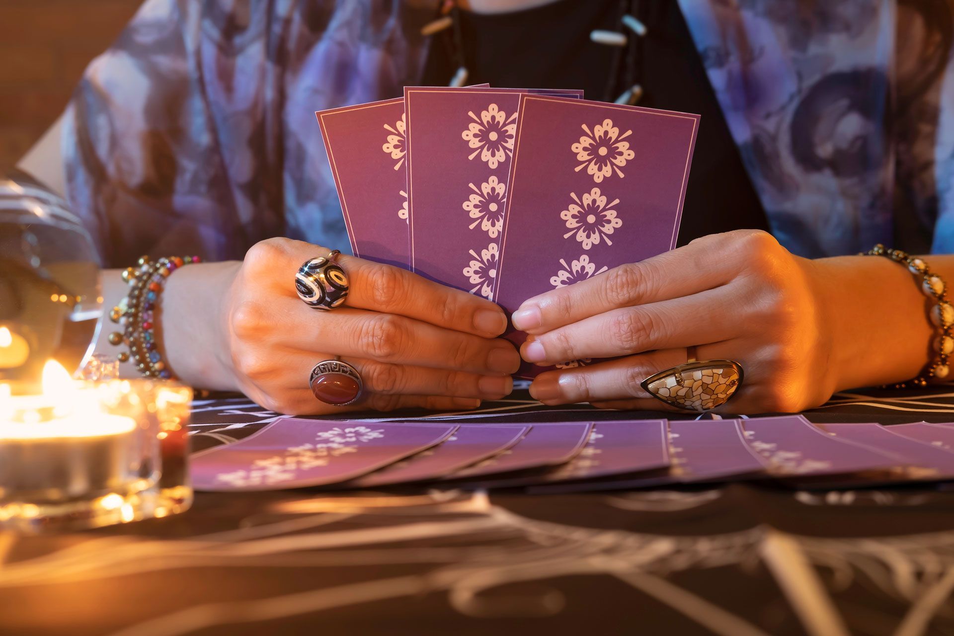 Hands holding tarot cards, purple backs with white flower pattern. Candles, table, jewelry.