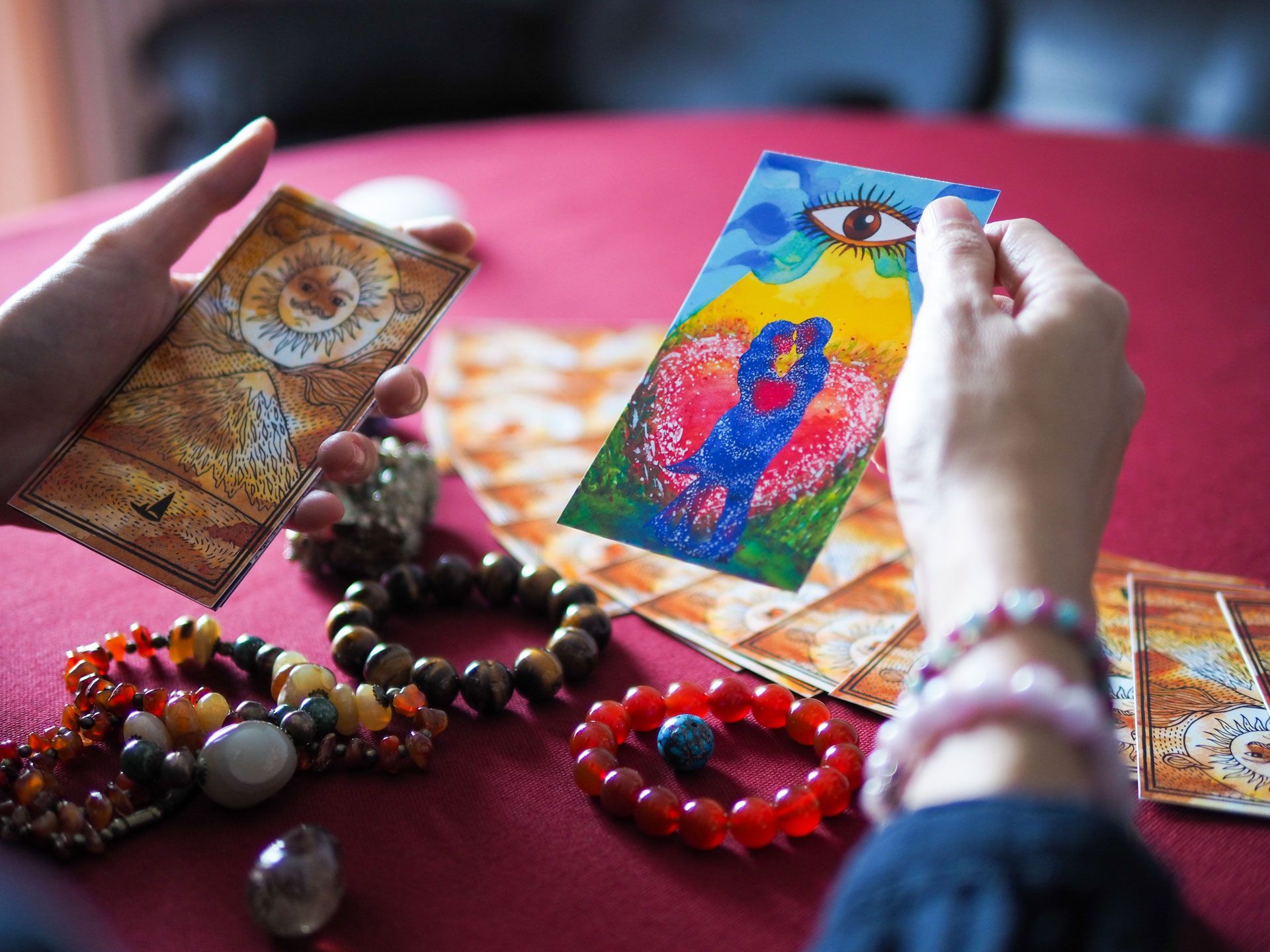 Hands holding tarot cards with colorful art, surrounded by bracelets and a red table.