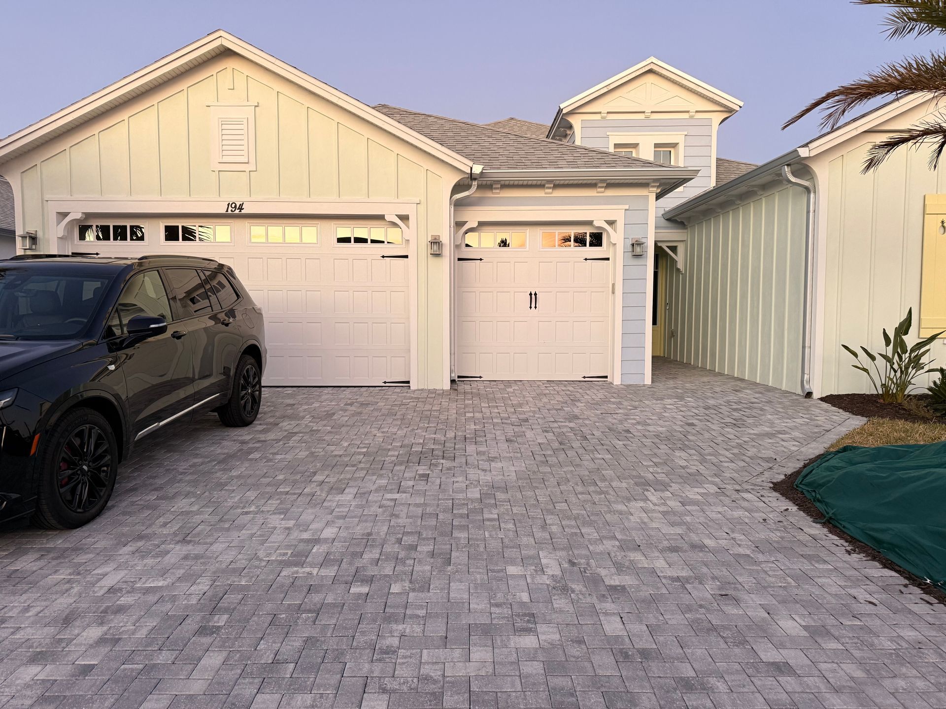 Gray brick driveway leading to a light-colored house with a black SUV parked in front.