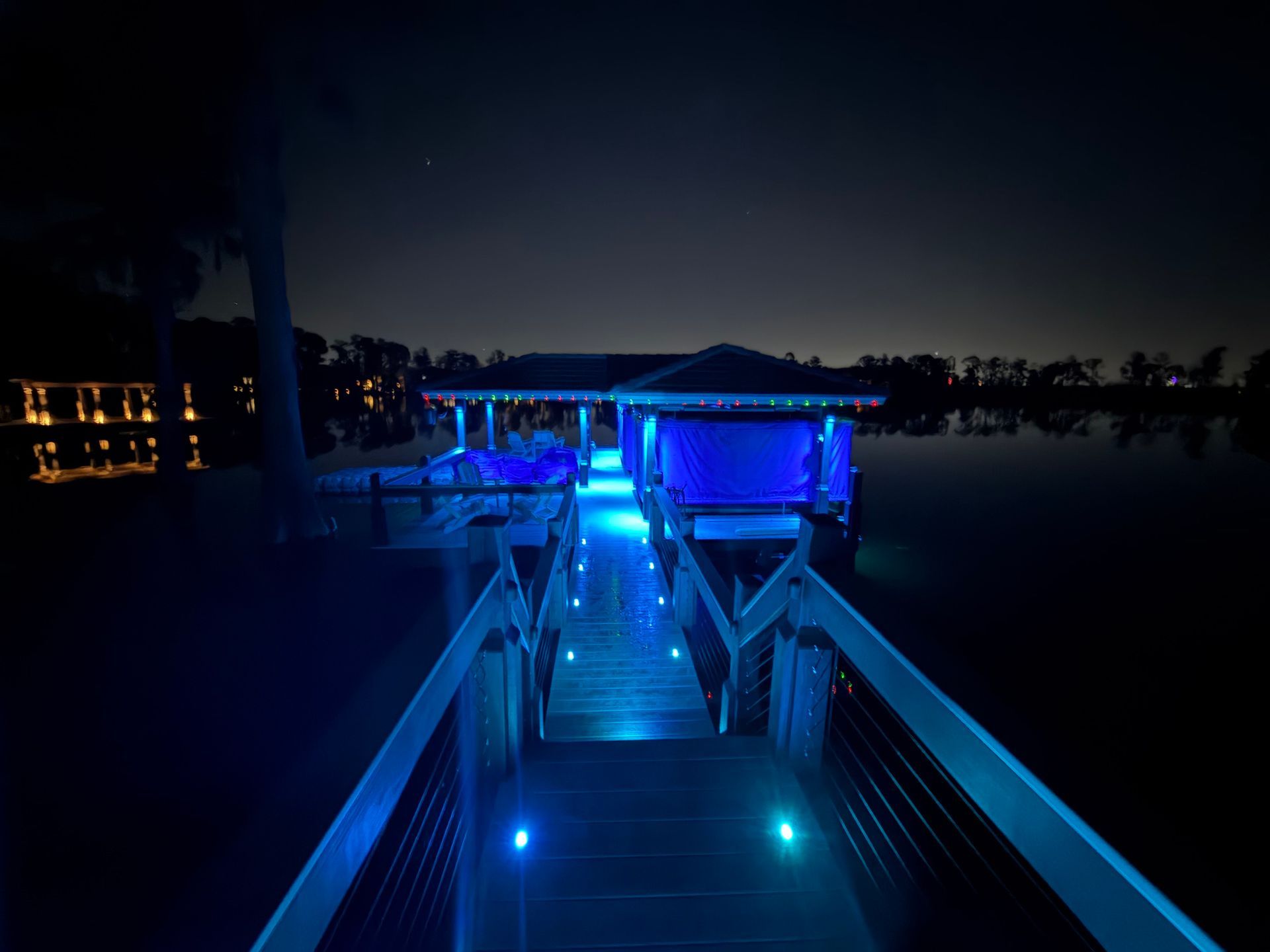 Wooden dock with blue lights leading to a covered structure over water at night.