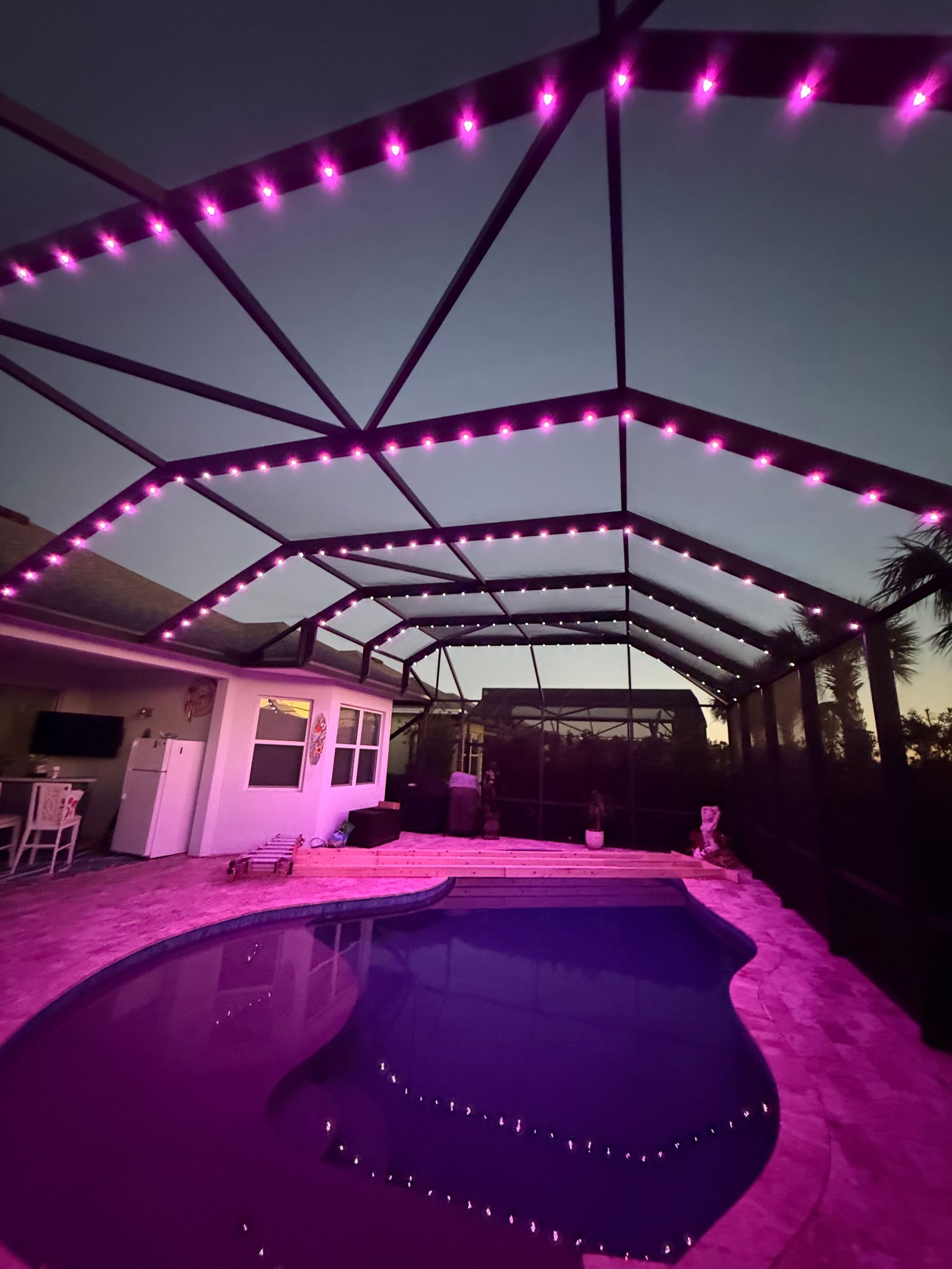 Pool area at dusk with pink lights illuminating the pool, enclosure, and house.