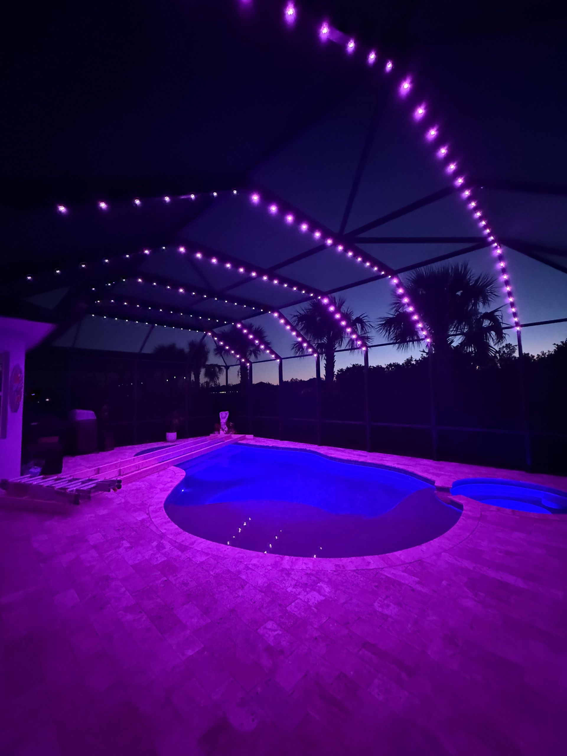 Purple string lights illuminate a pool area at dusk. The pool and patio glow with purple light.