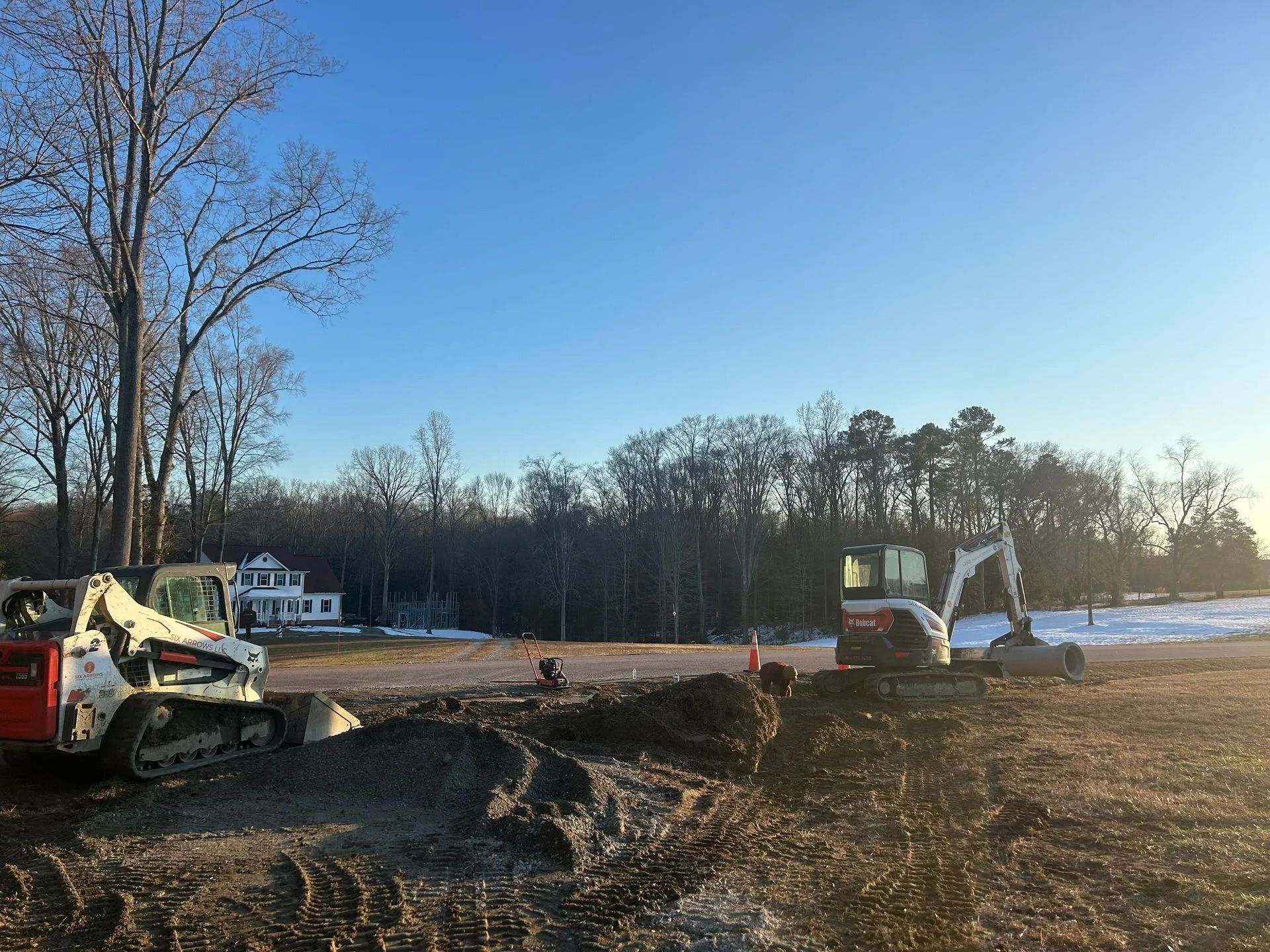 Two construction vehicles sit on a dirt lot under a bright blue sky, with trees and a distant building in the background.