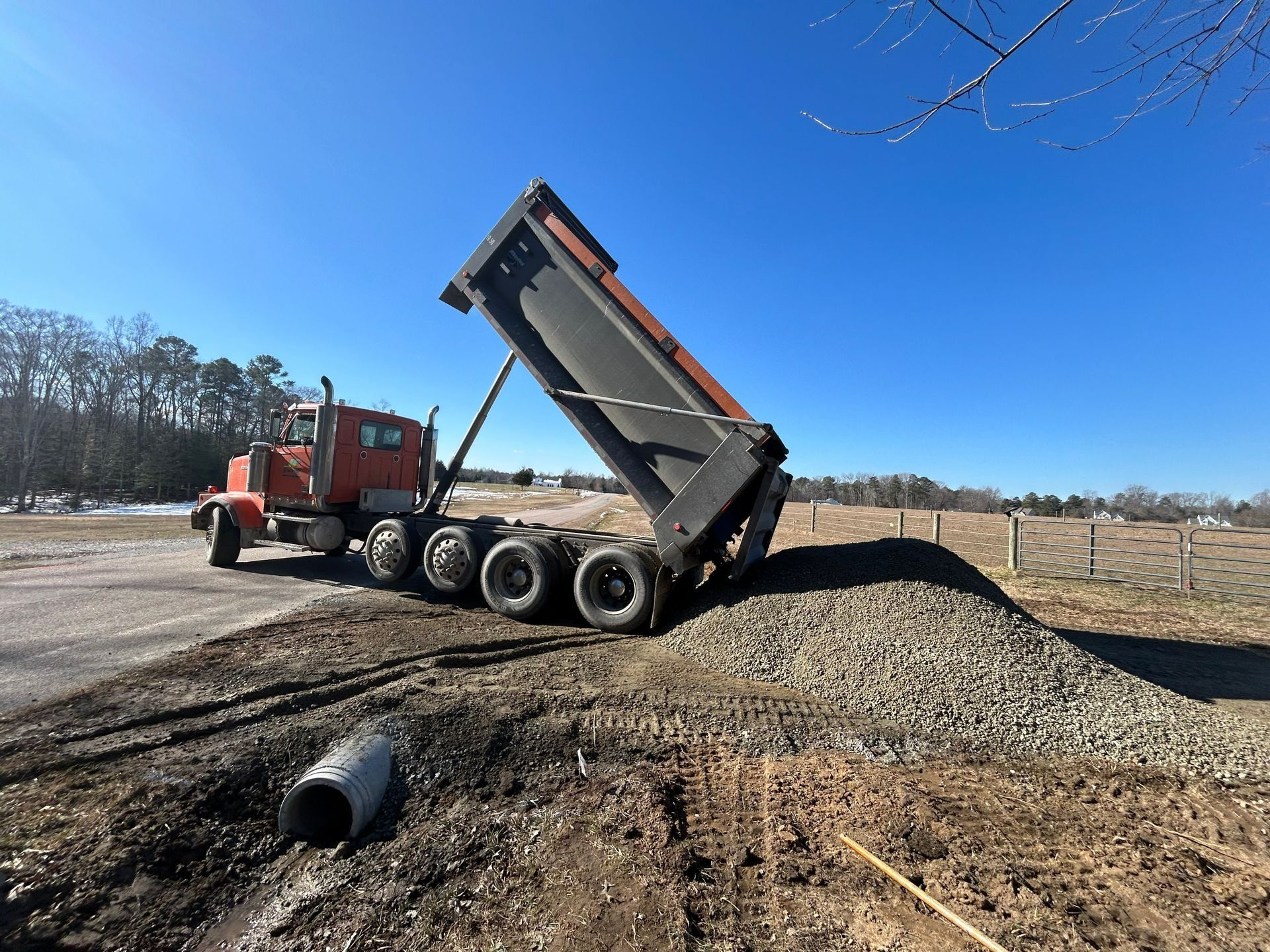 An orange dump truck parked on a dirt lot is raised to deposit a pile of gravel onto the ground under a clear blue sky.
