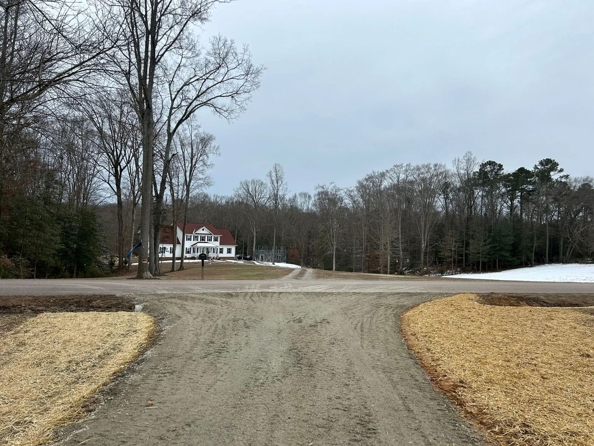 A gravel driveway leads toward a white, two-story house nestled among bare trees under an overcast sky.