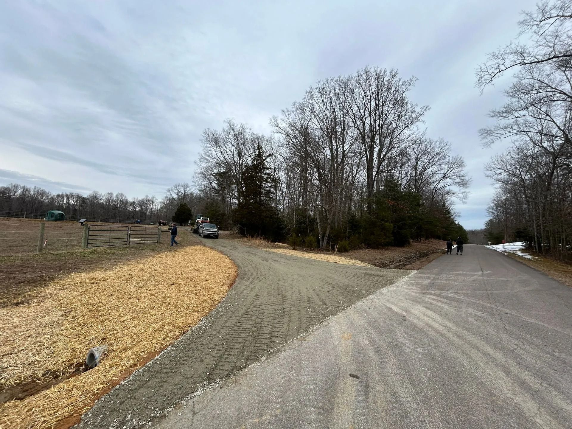 A gravel driveway meets an asphalt road near a field, with a patch of light-colored wood chips in the foreground.
