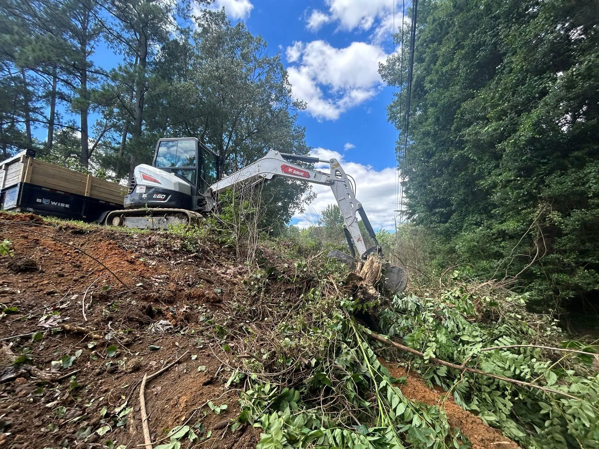 A white excavator clears brush and debris on a hillside next to a trailer under a blue, cloudy sky.