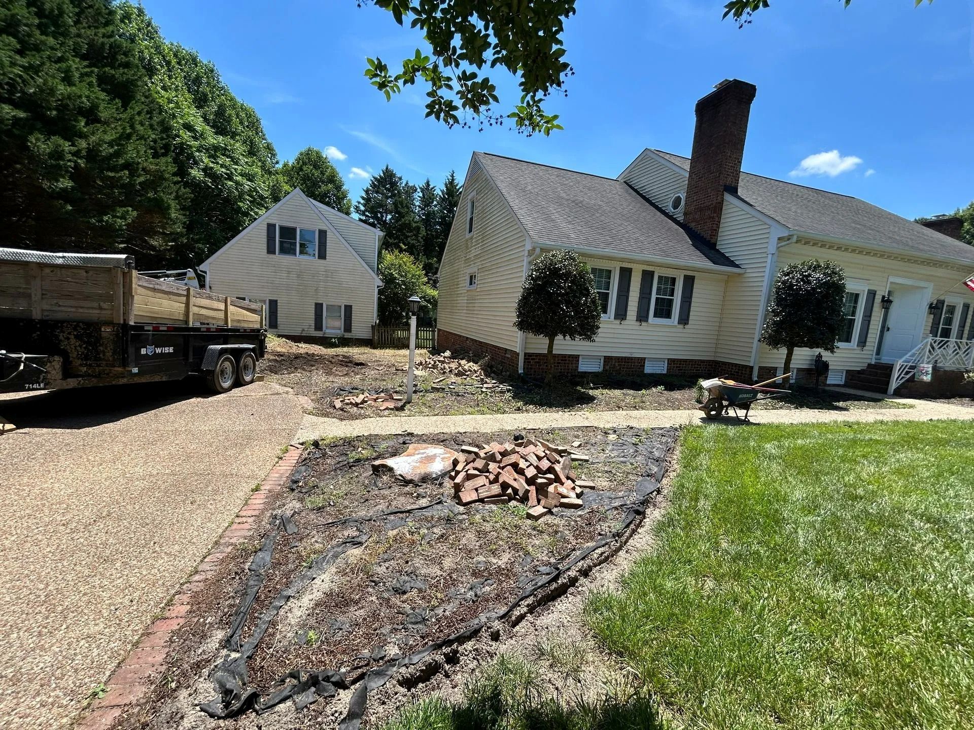 A light-colored house with a brick chimney stands next to a driveway with a trailer, near a cleared dirt landscape patch.