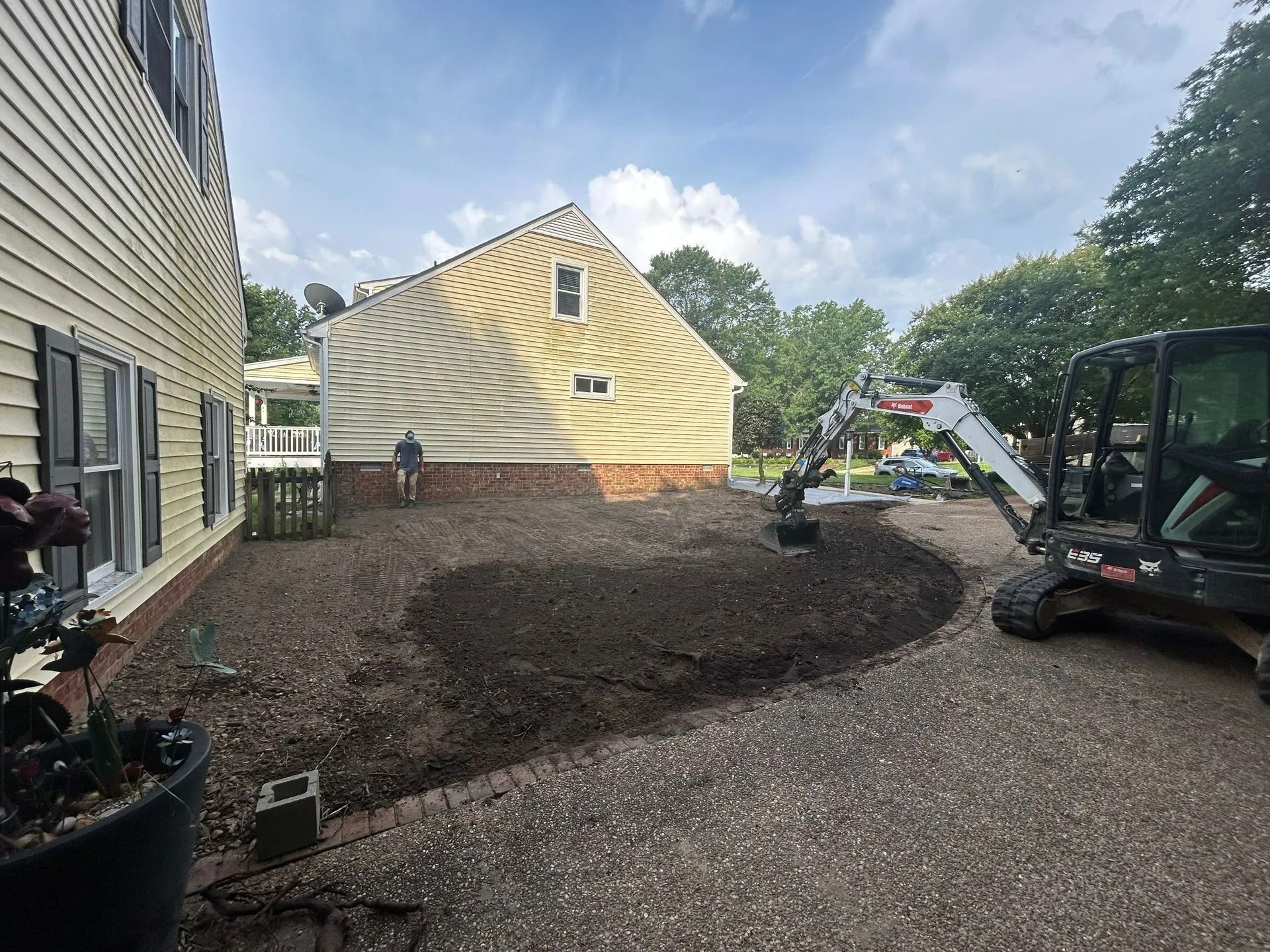 A compact excavator sits in a residential yard as a person works near a light-colored house with a brick foundation.