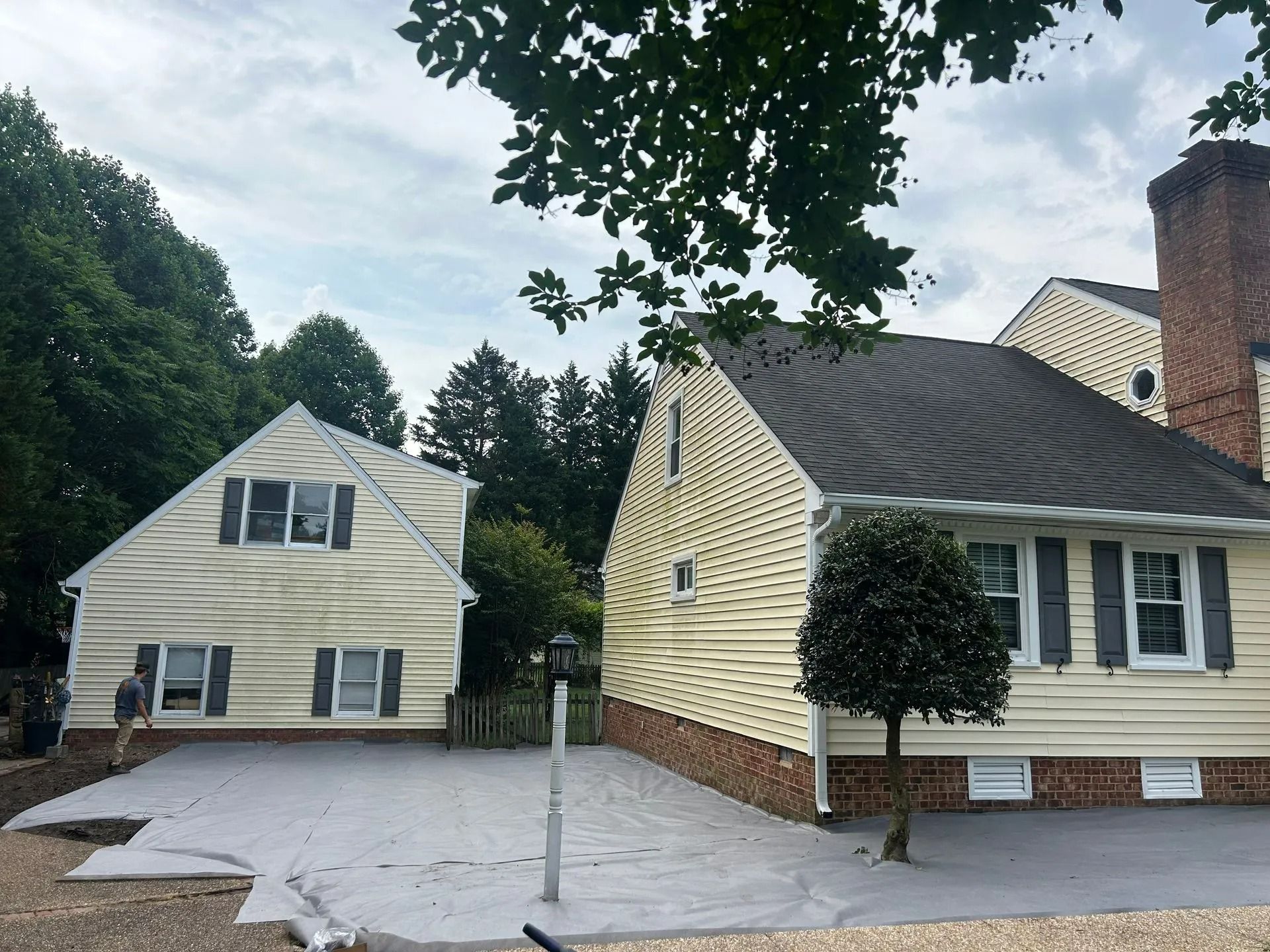 Two light-yellow houses with dark shutters sit beside a grey patio area under a cloudy sky.