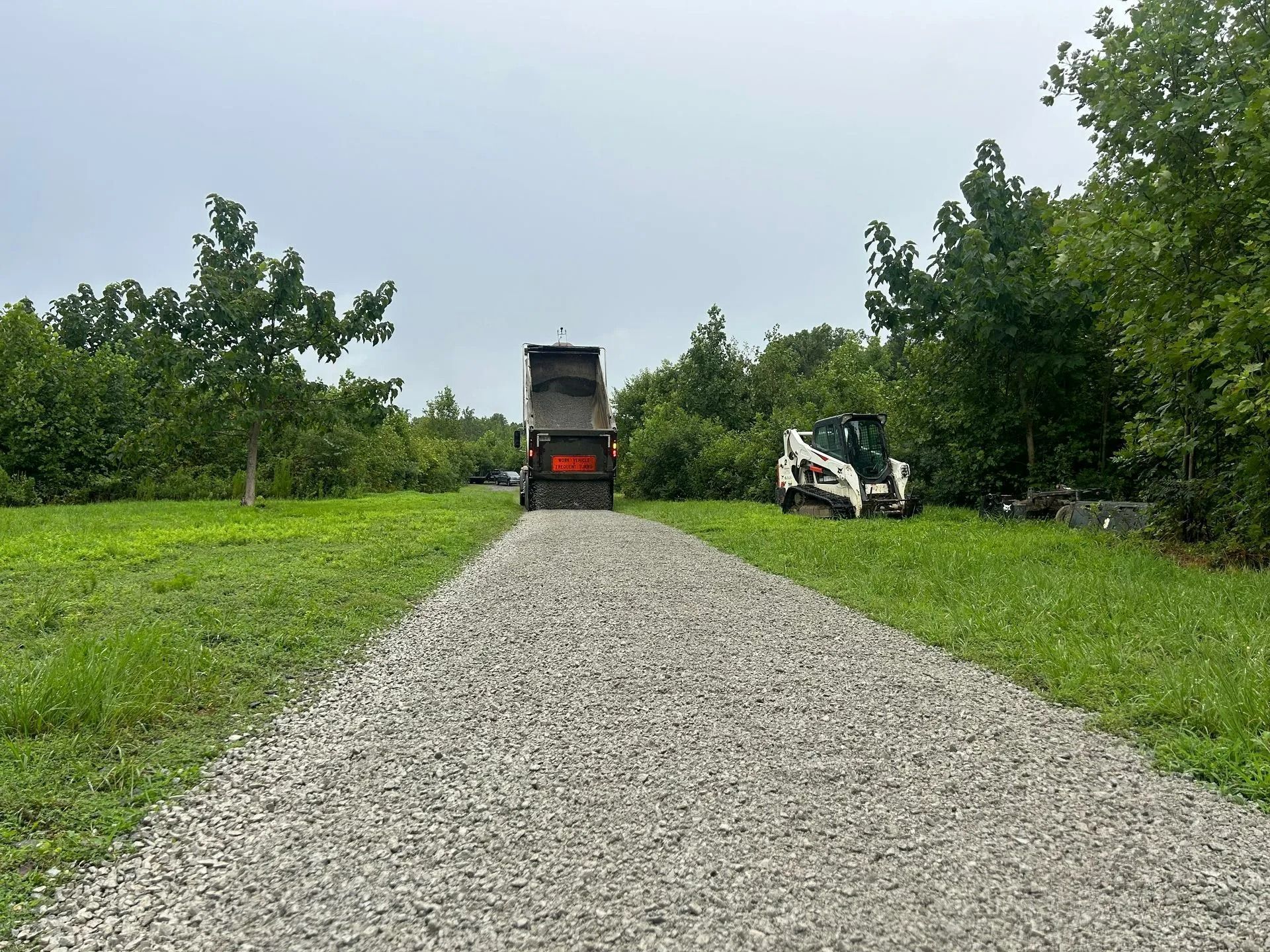 A dump truck delivers gravel to a new stone path in a green, grassy field with a white skid-steer parked nearby.
