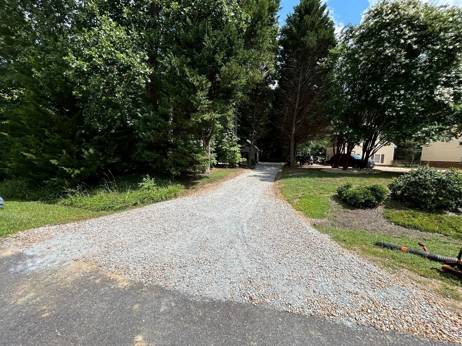 A gravel driveway transitions from asphalt, leading toward trees and a house in a sunny, green outdoor setting.