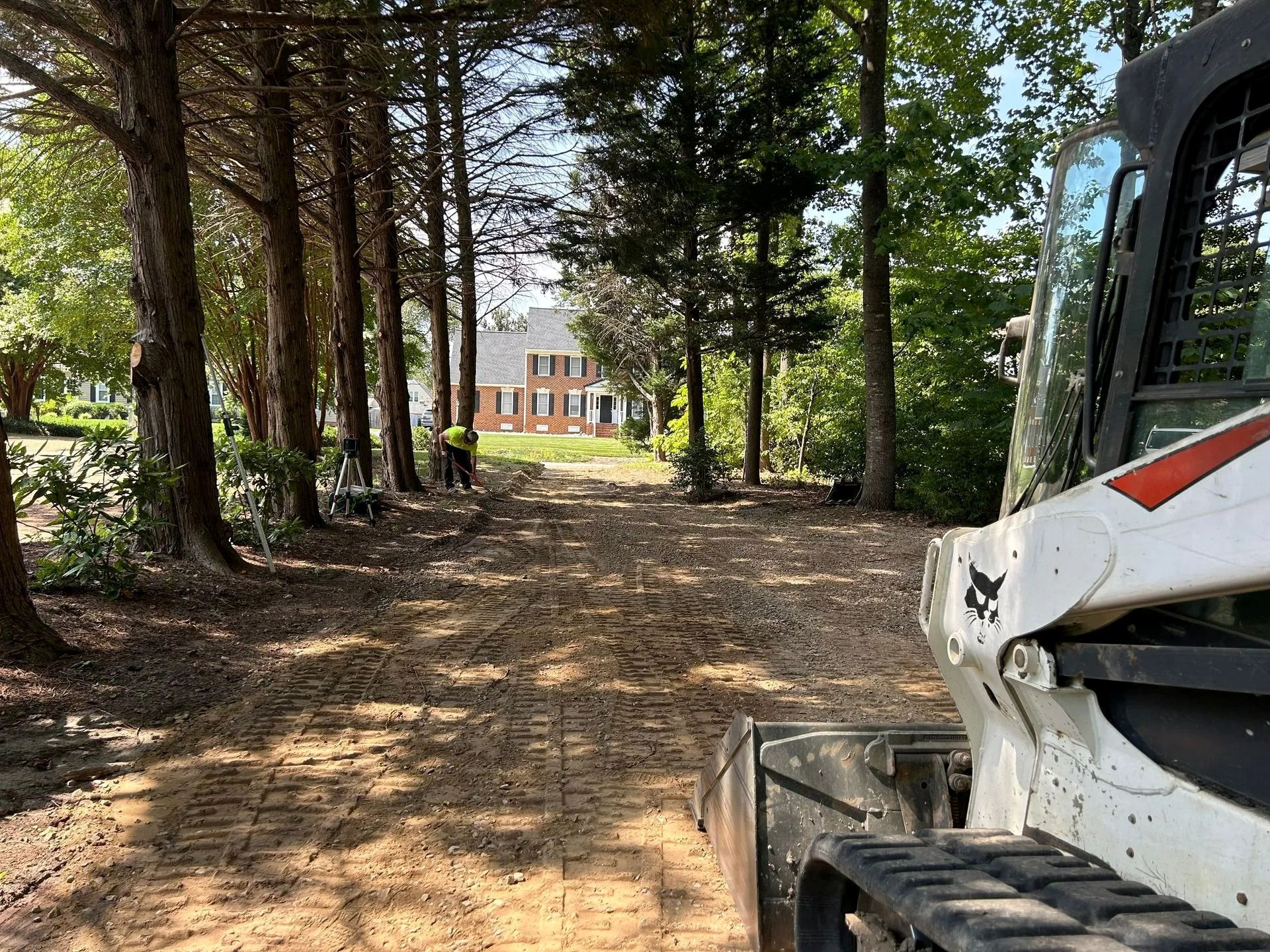 A skid steer loader sits on a dirt path in a wooded area, with a house visible in the distance under a blue sky.
