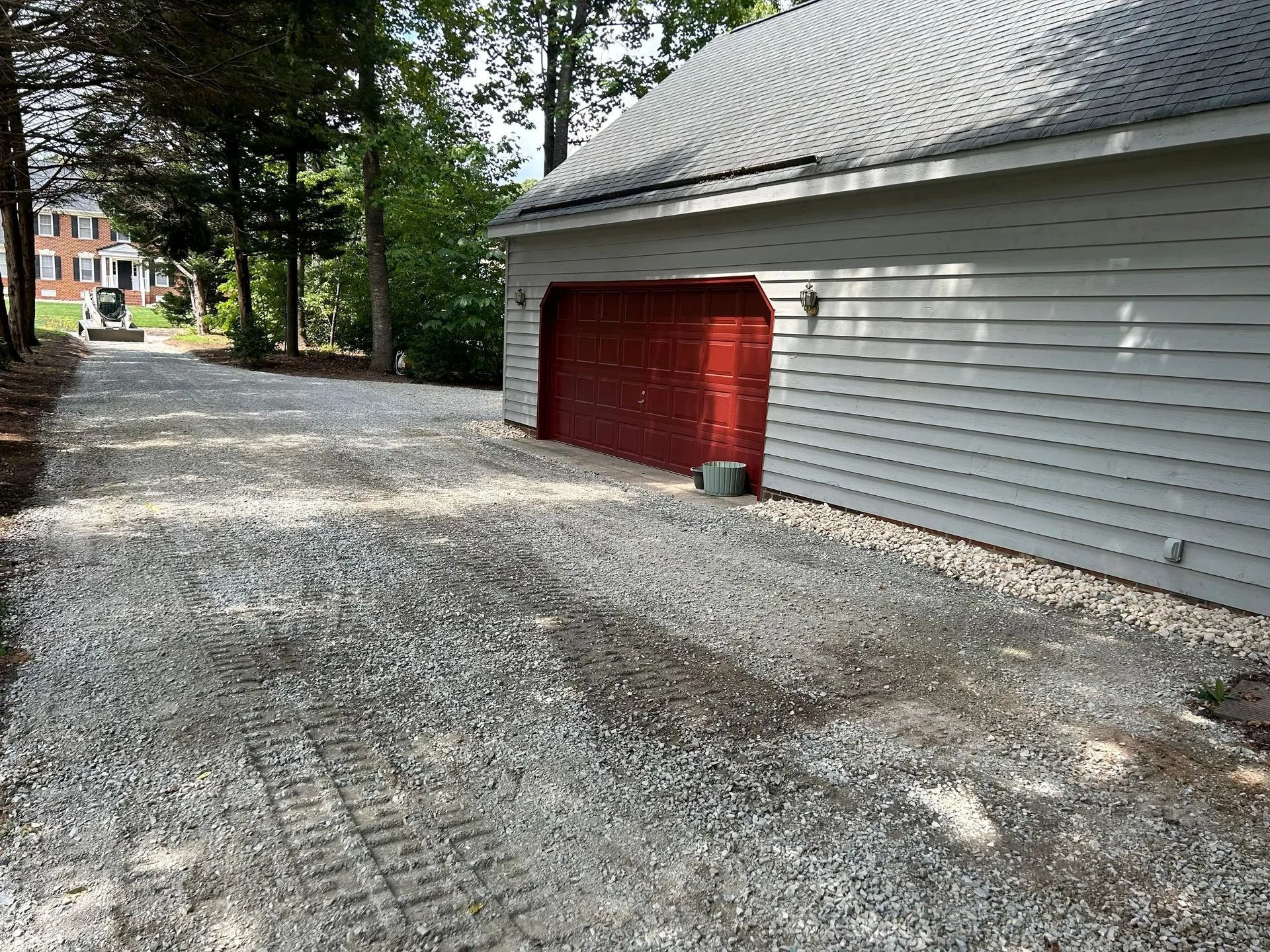 A gravel driveway leads to a light-colored garage with a dark red door, surrounded by trees on a sunny day.