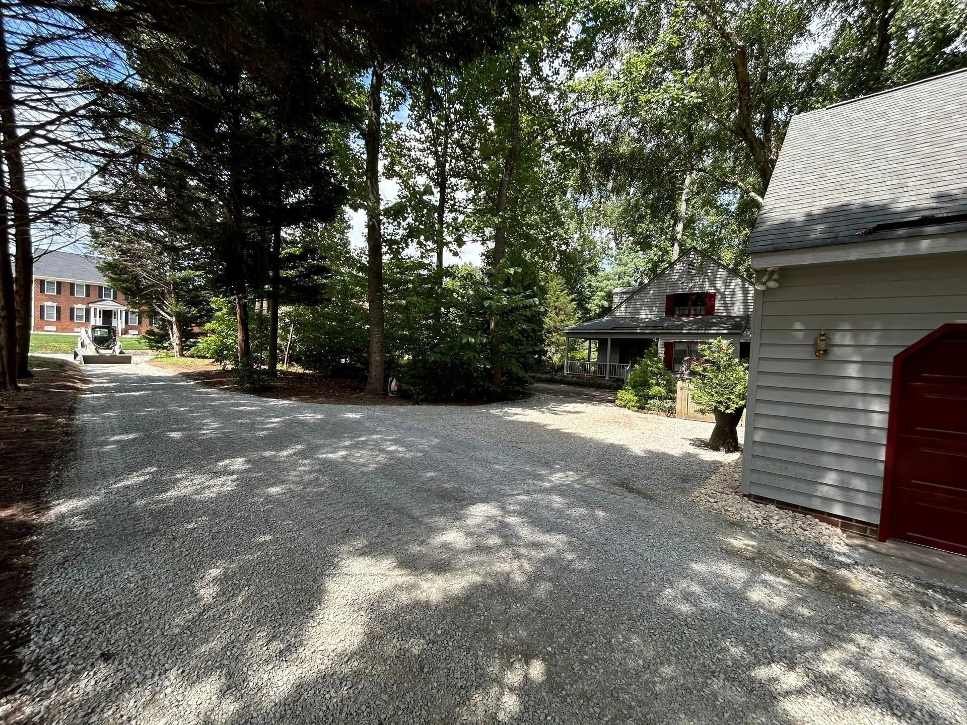 A gravel driveway leads toward a house and a garage with a red door, surrounded by green trees on a sunny day.
