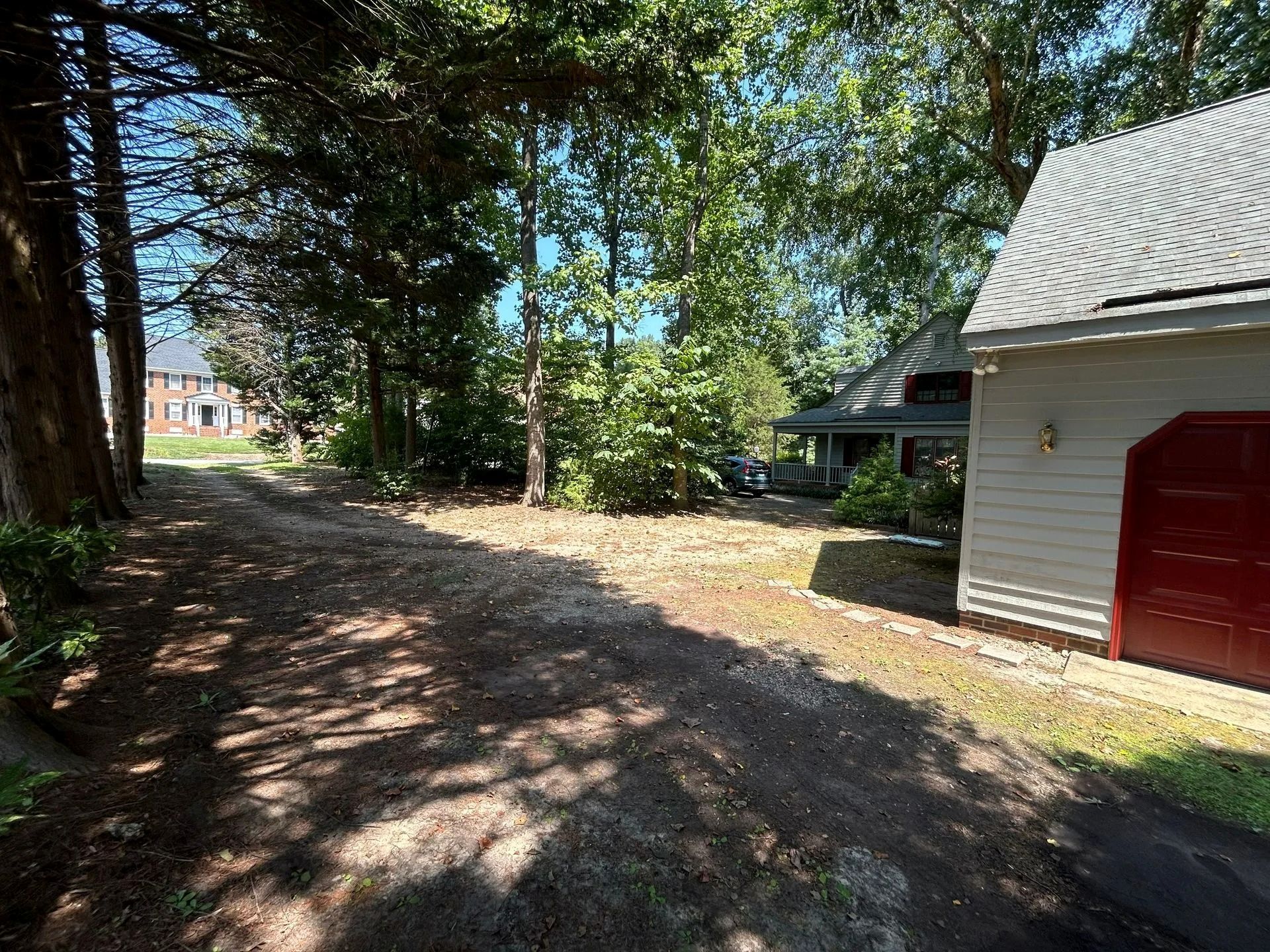 A dirt path leads through a shaded yard with trees and bushes beside a house with a red garage door.