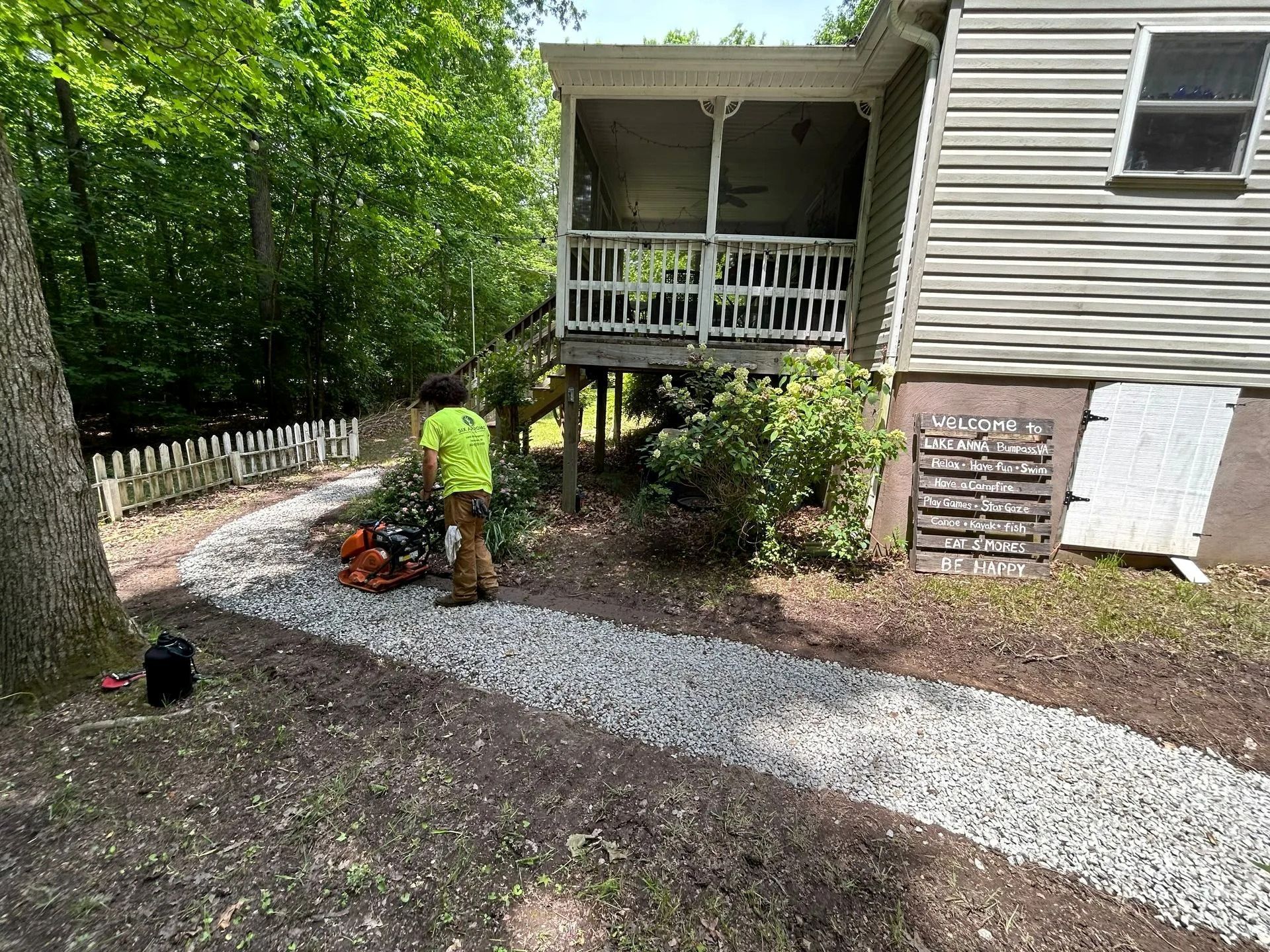 A person in a neon yellow shirt works on a gray gravel path next to a house with a wooden porch and a welcome sign.