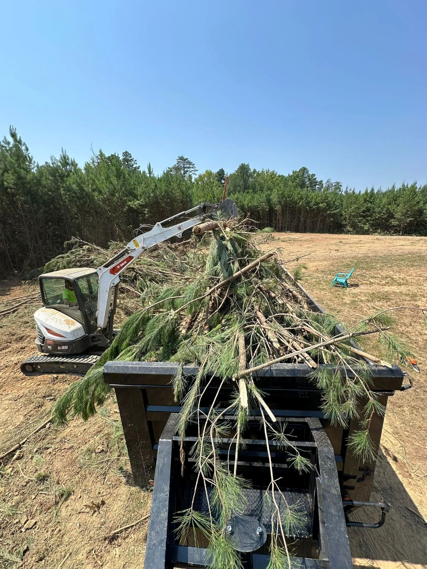 A white Bobcat excavator lifts branches into a large metal trailer on a sunny, cleared plot of land.