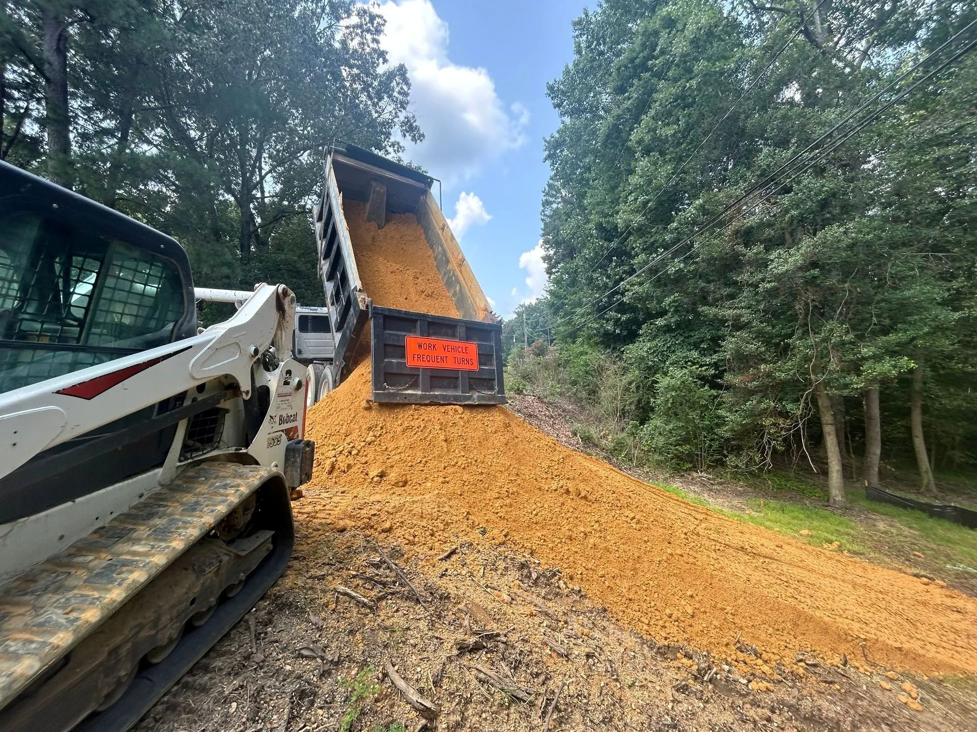 A dump truck pours a pile of orange dirt onto a path near a forest, with a white track loader parked nearby.