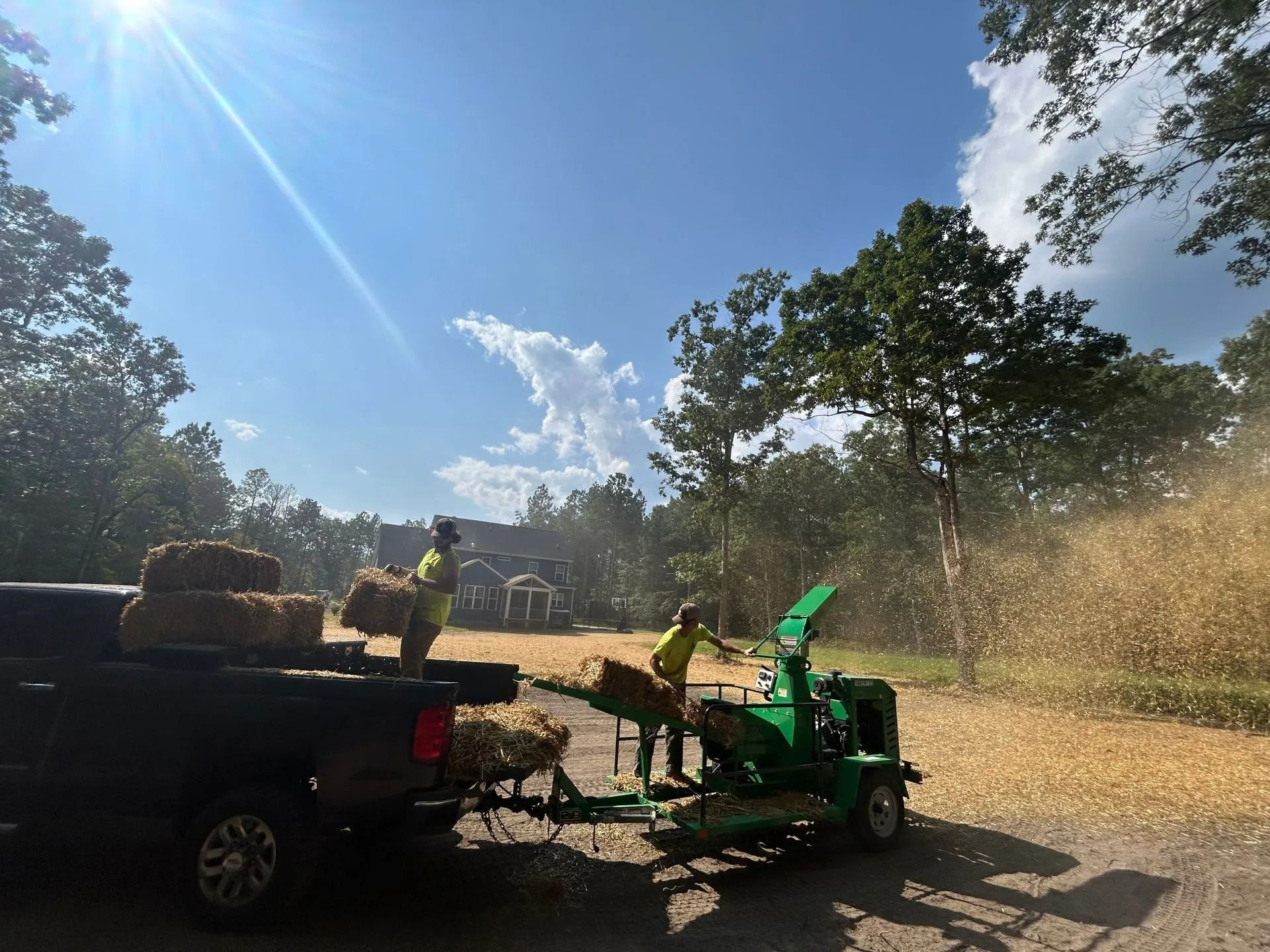 Two people in yellow shirts feed straw into a green wood chipper attached to a truck, creating a cloud of straw debris.