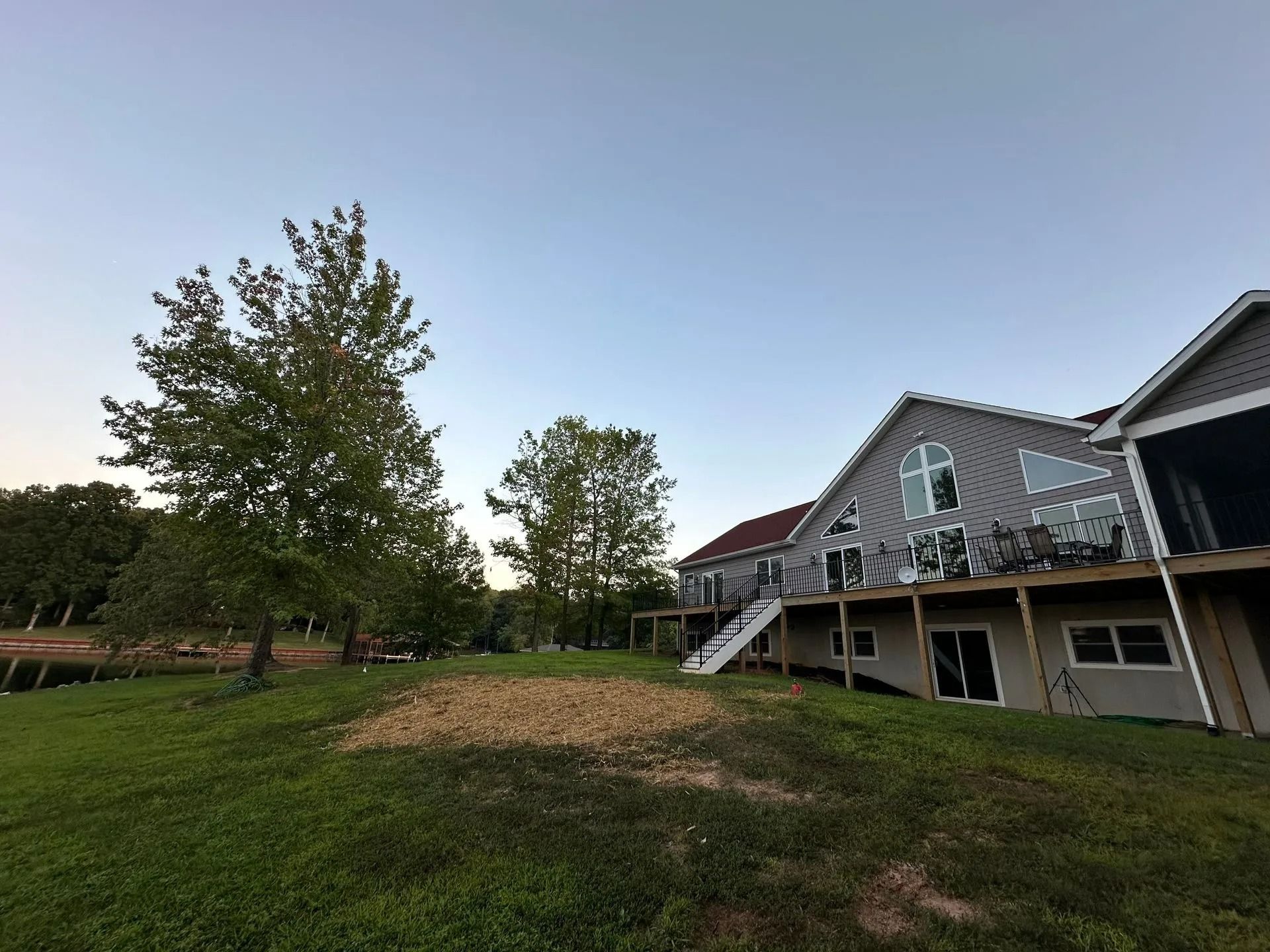 A wide-angle view of a grey multi-story house with a wooden deck, set on a grassy hill near trees at sunset.
