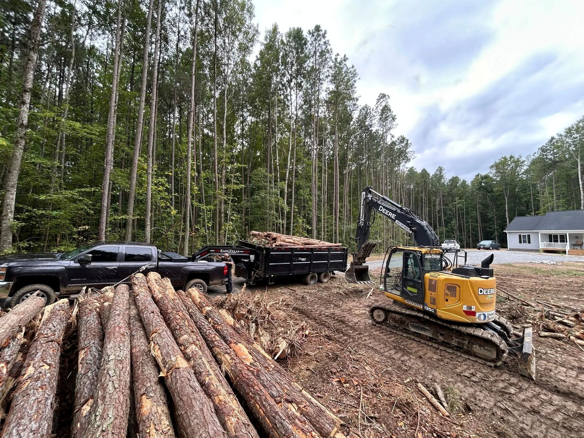 A yellow excavator loads logs into a large black trailer parked near a forest edge and a residential house.