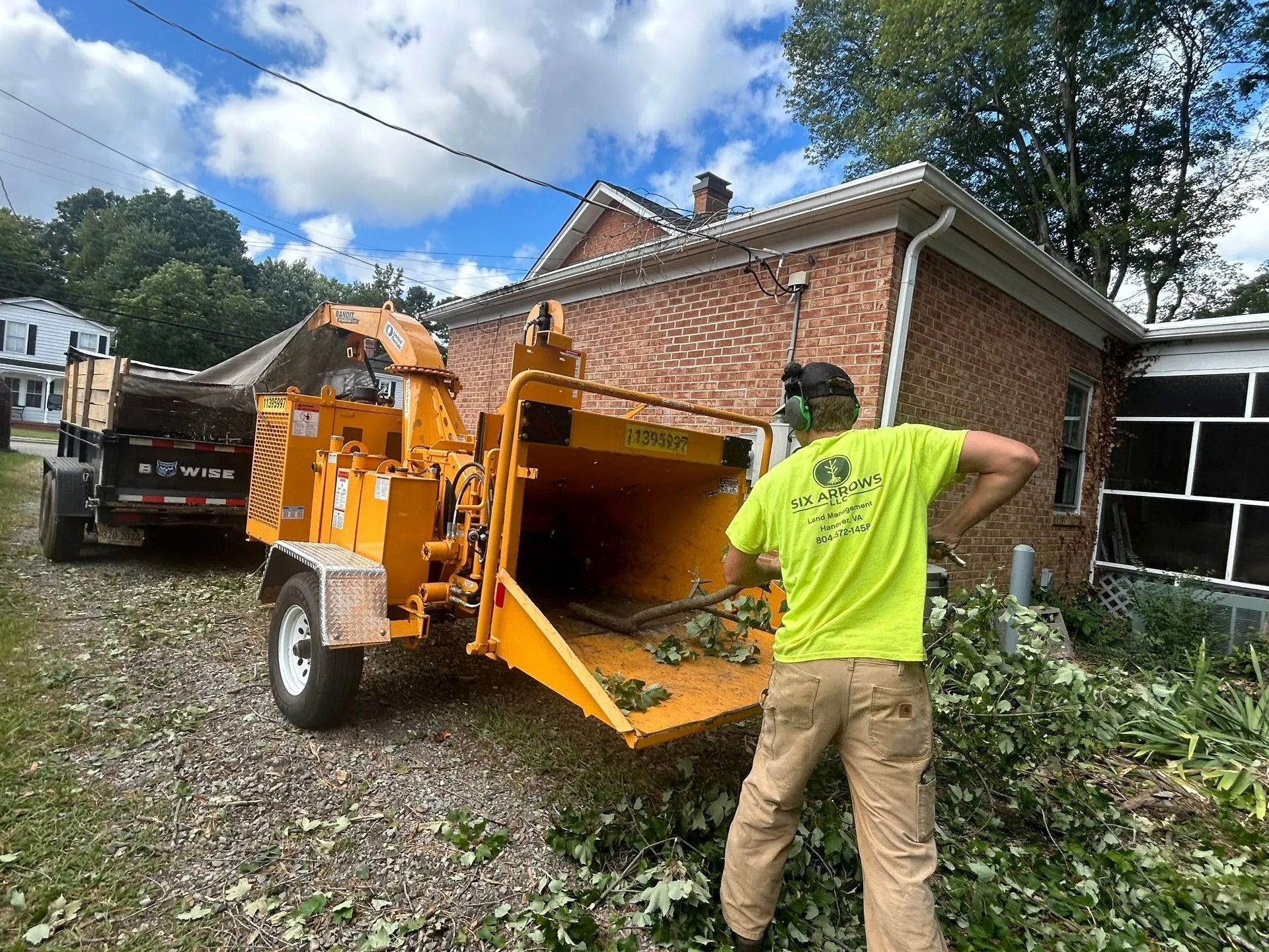 A worker in a neon yellow shirt feeds tree branches into a large, yellow industrial wood chipper next to a brick house.